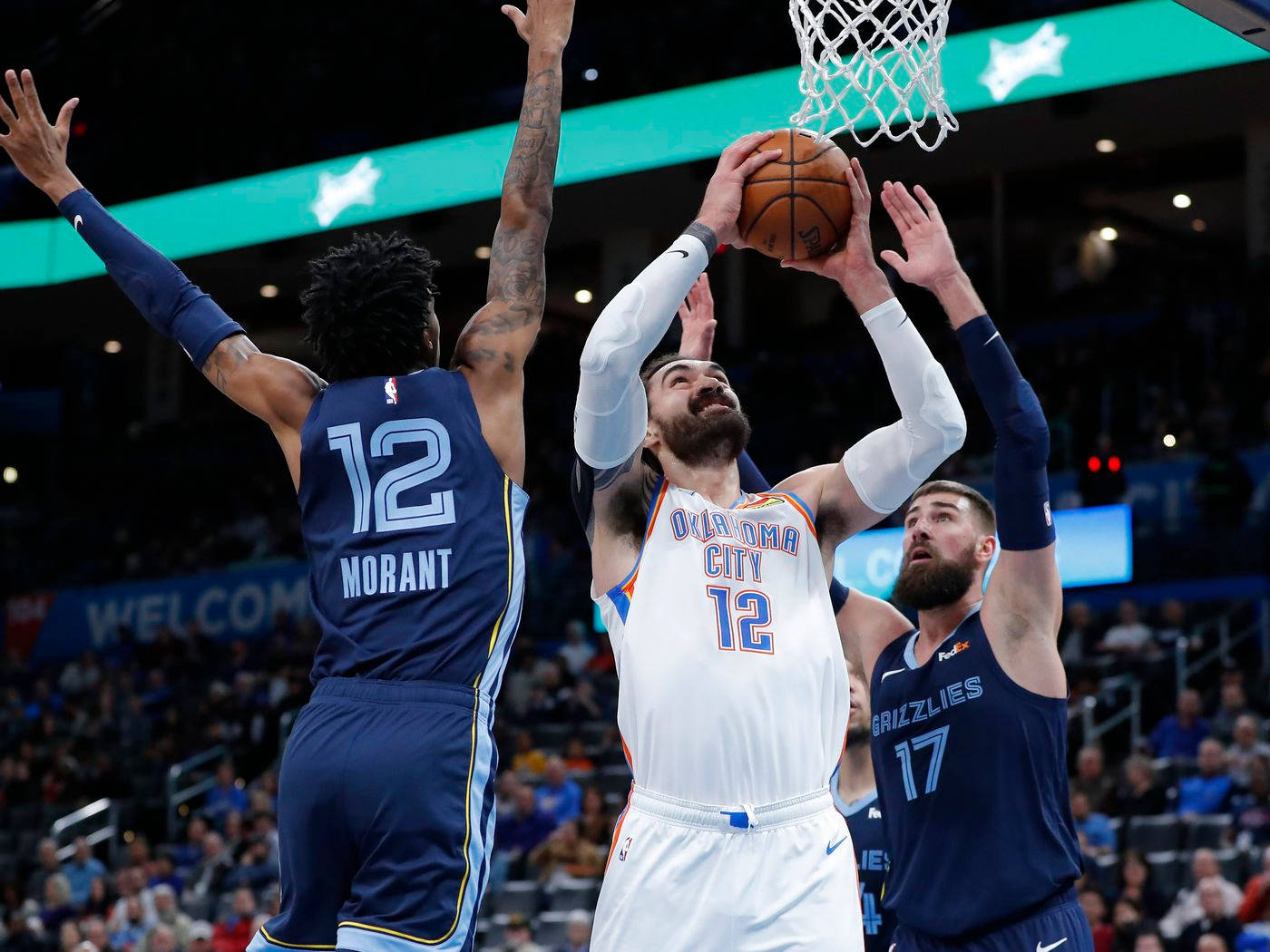 Steven Adams 桌布 壁紙 — 一名身穿白色奧克拉荷馬城球衣的籃球運動員跳起投籃，兩名身穿藍色球衣的防守球員在明亮的場館燈光下干擾投籃。