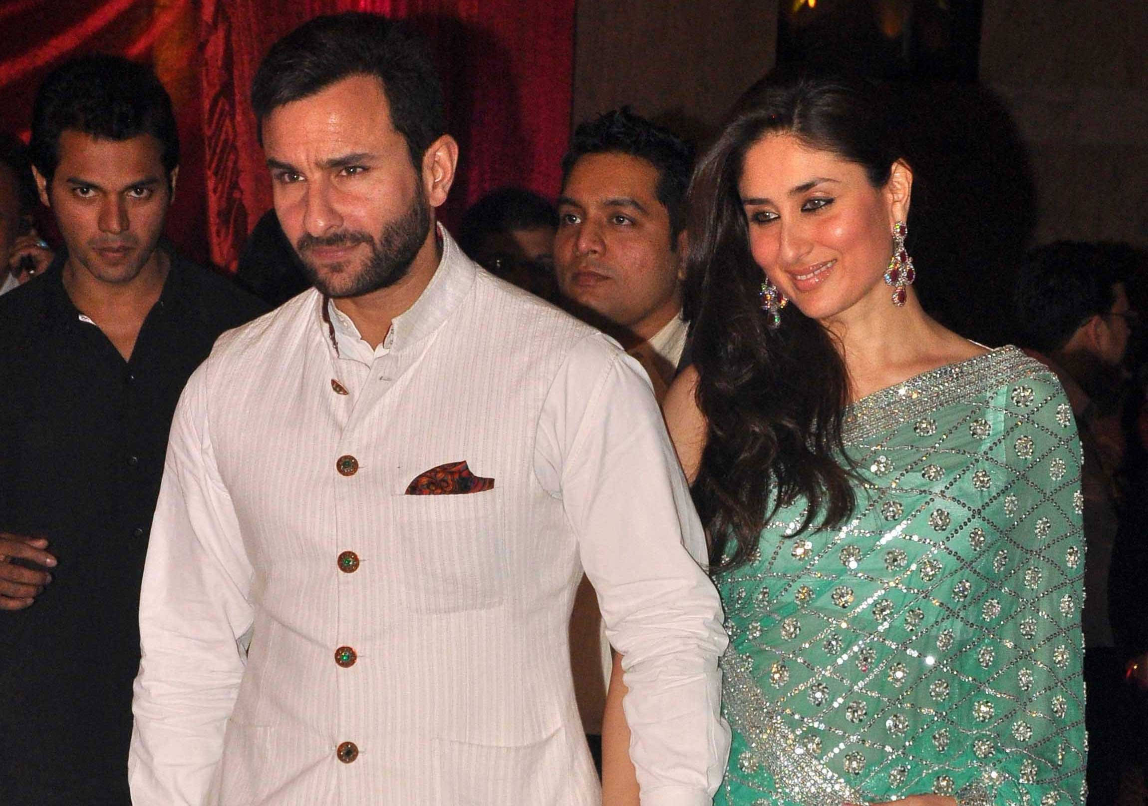 Saif Ali Khan 桌布 壁紙 — 一名身穿白色庫爾塔的男子和一名身穿青綠色裝飾紗麗的女子，在燈光昏暗、身後有紅色帷幕的活動中站在一起。