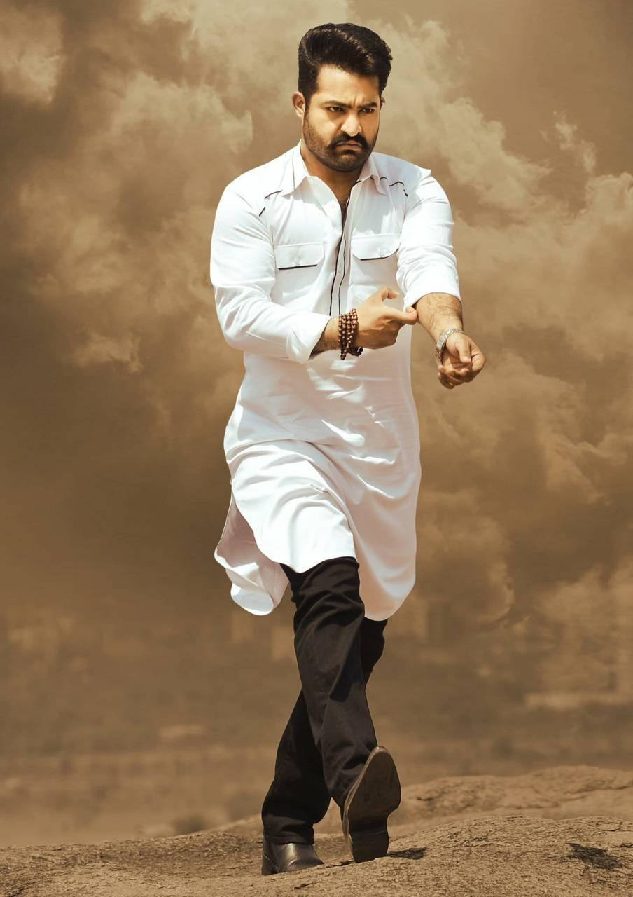 jr ntr 桌布 壁紙 — 一名身穿白色庫爾塔的男子在戲劇性的多雲天空下自信地走過塵土飛揚的地形。