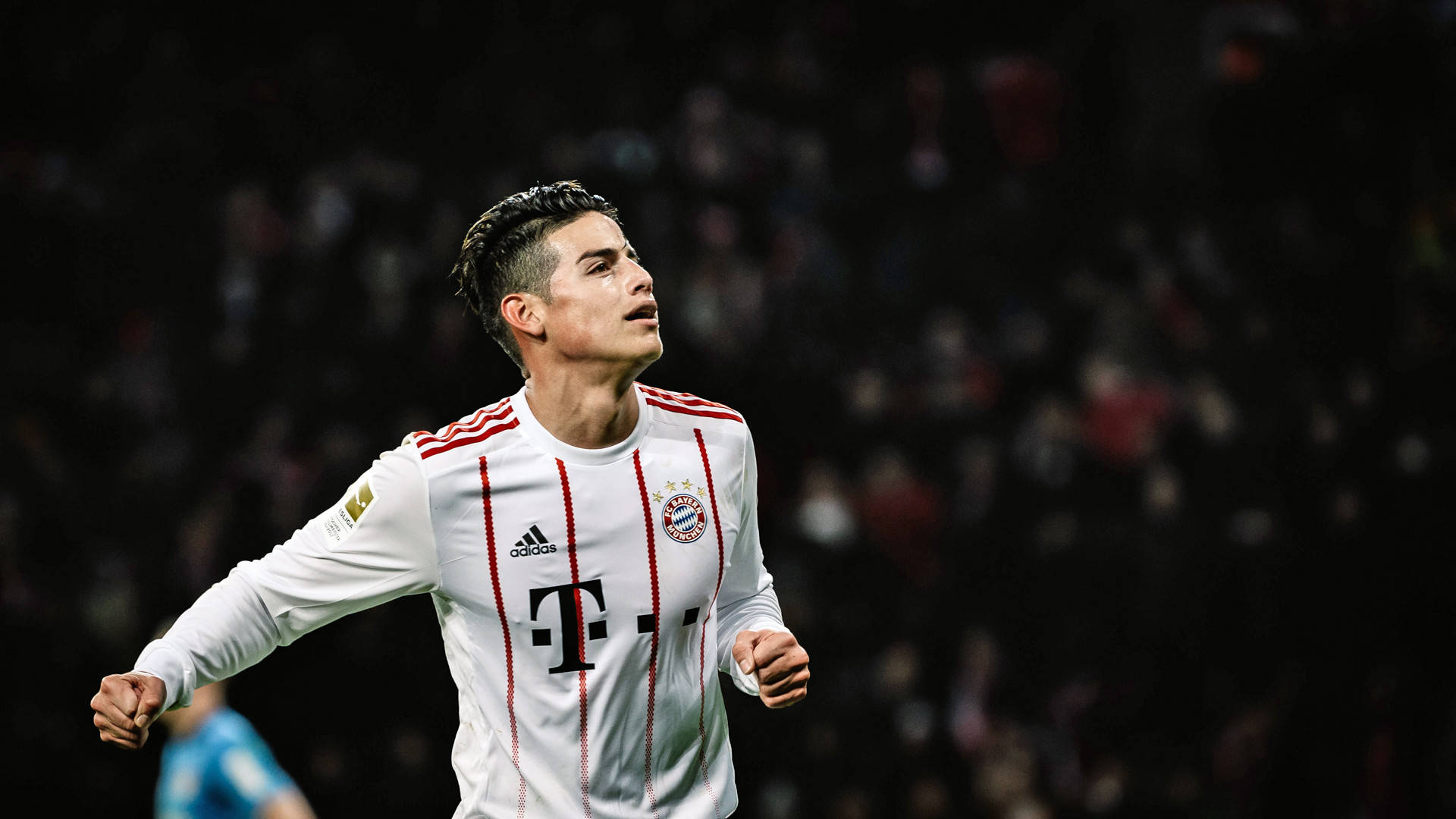 James Rodriguez 桌布 壁紙 — 一名身穿白色拜仁慕尼黑球衣的足球運動員在球場燈光下握緊拳頭慶祝。