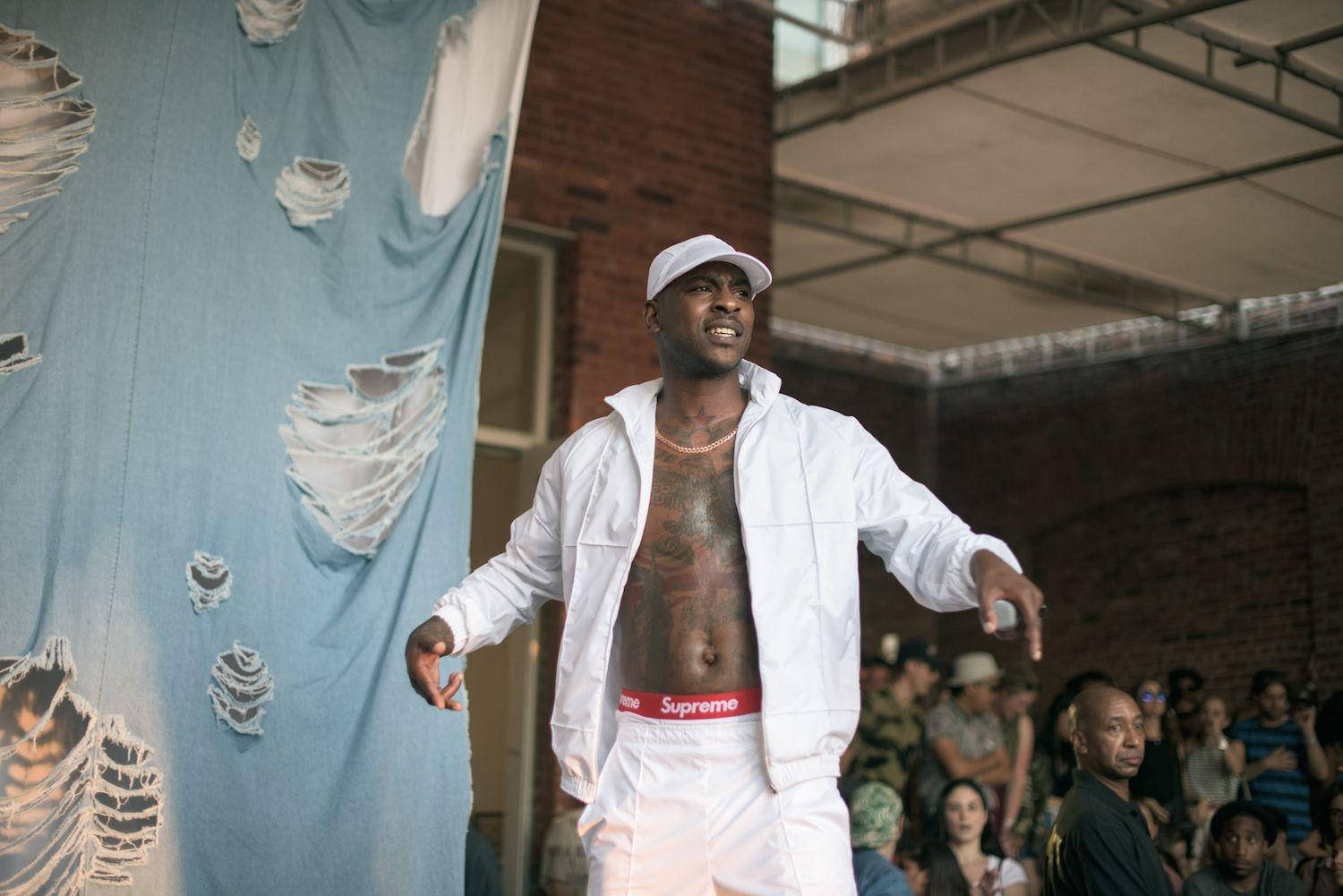 Skepta 桌布 壁紙 — 一名身穿白色服裝和帽子的男子在舞台上充滿活力地表演，背景是撕裂的藍色背景和磚牆。