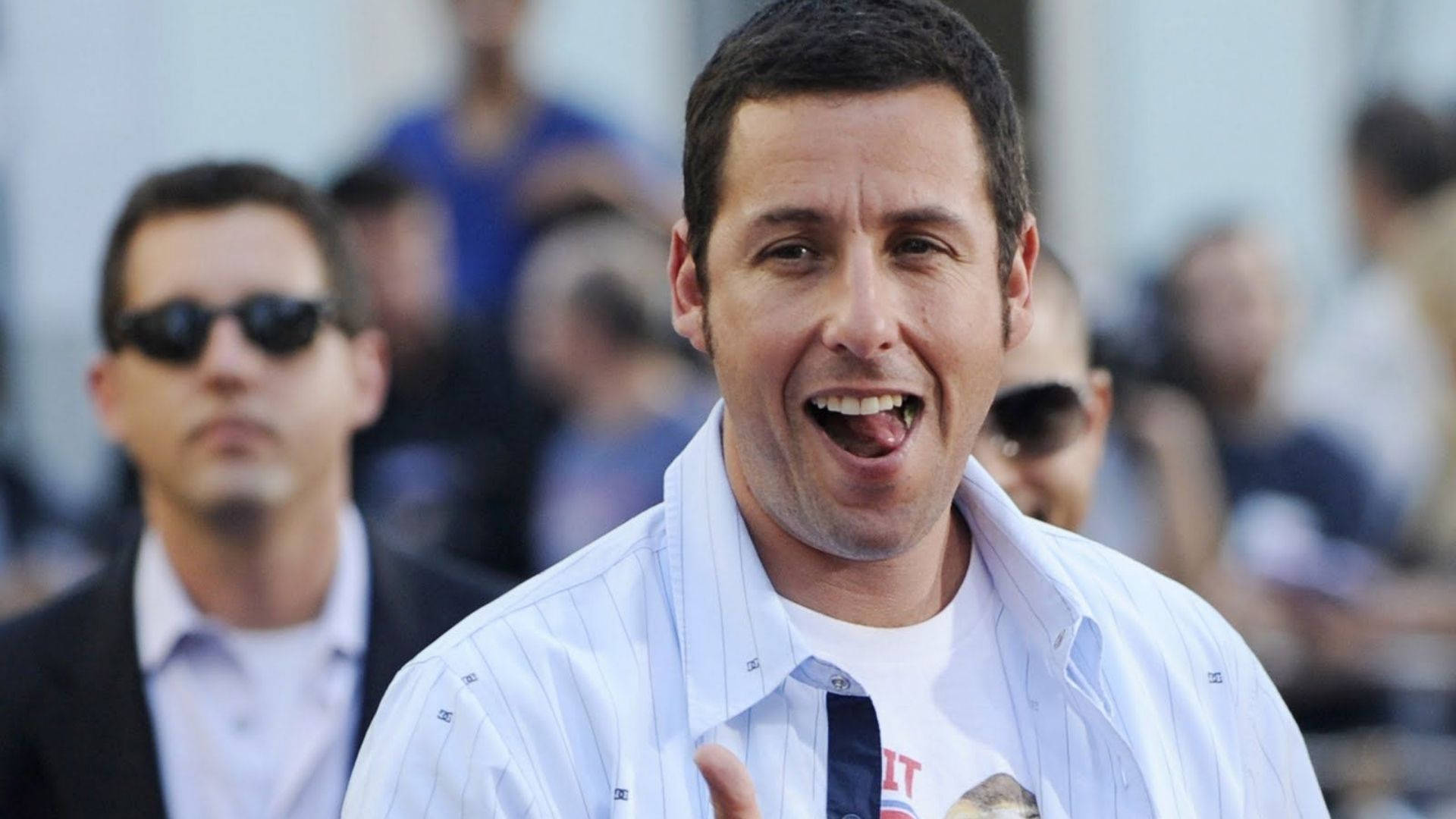Adam Sandler 桌布 壁紙 — 一名身穿白色條紋襯衫的男子在模糊的人群中張開嘴大笑，伸出舌頭。