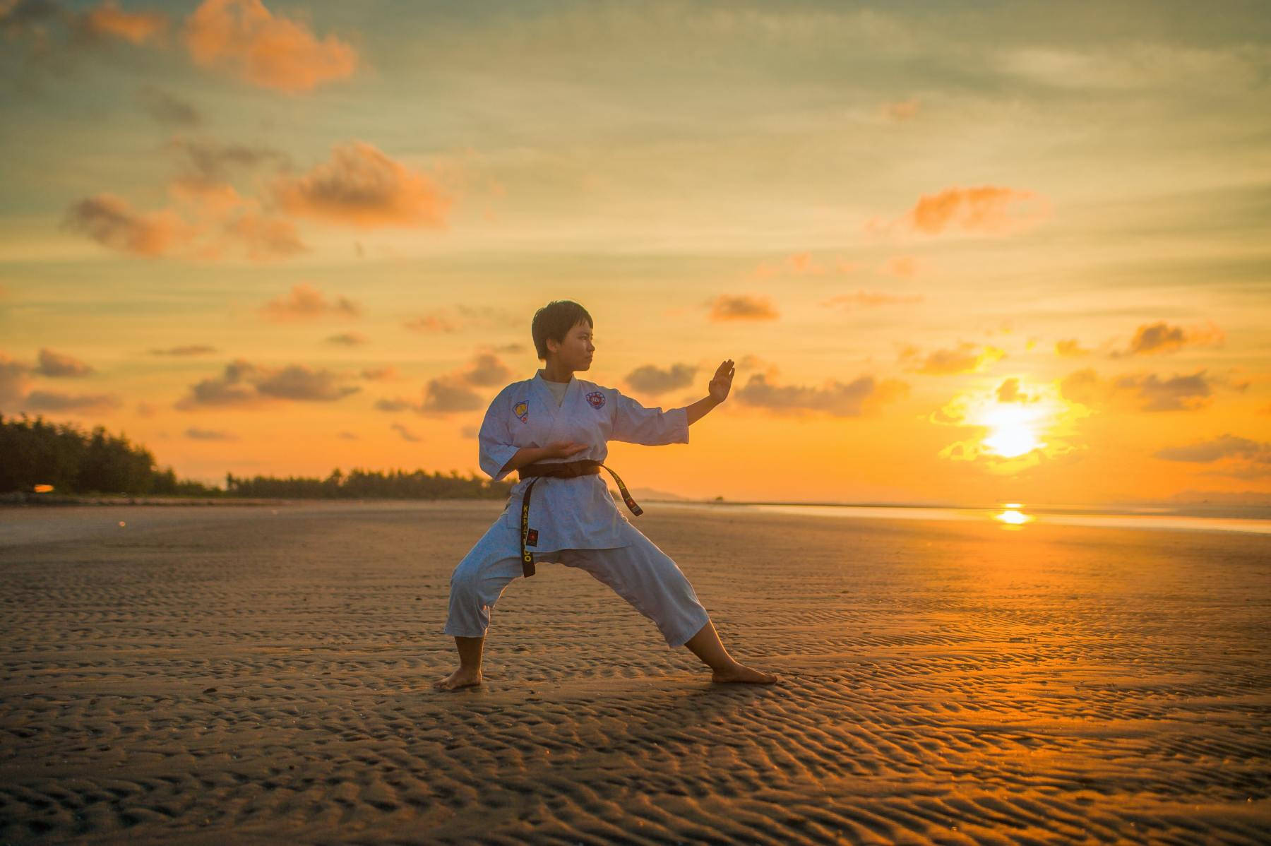 martial arts 桌布 壁紙 — 一名身穿白色武術制服的人在金色日落和溫暖發光的雲彩下，於沙灘上練習姿勢。