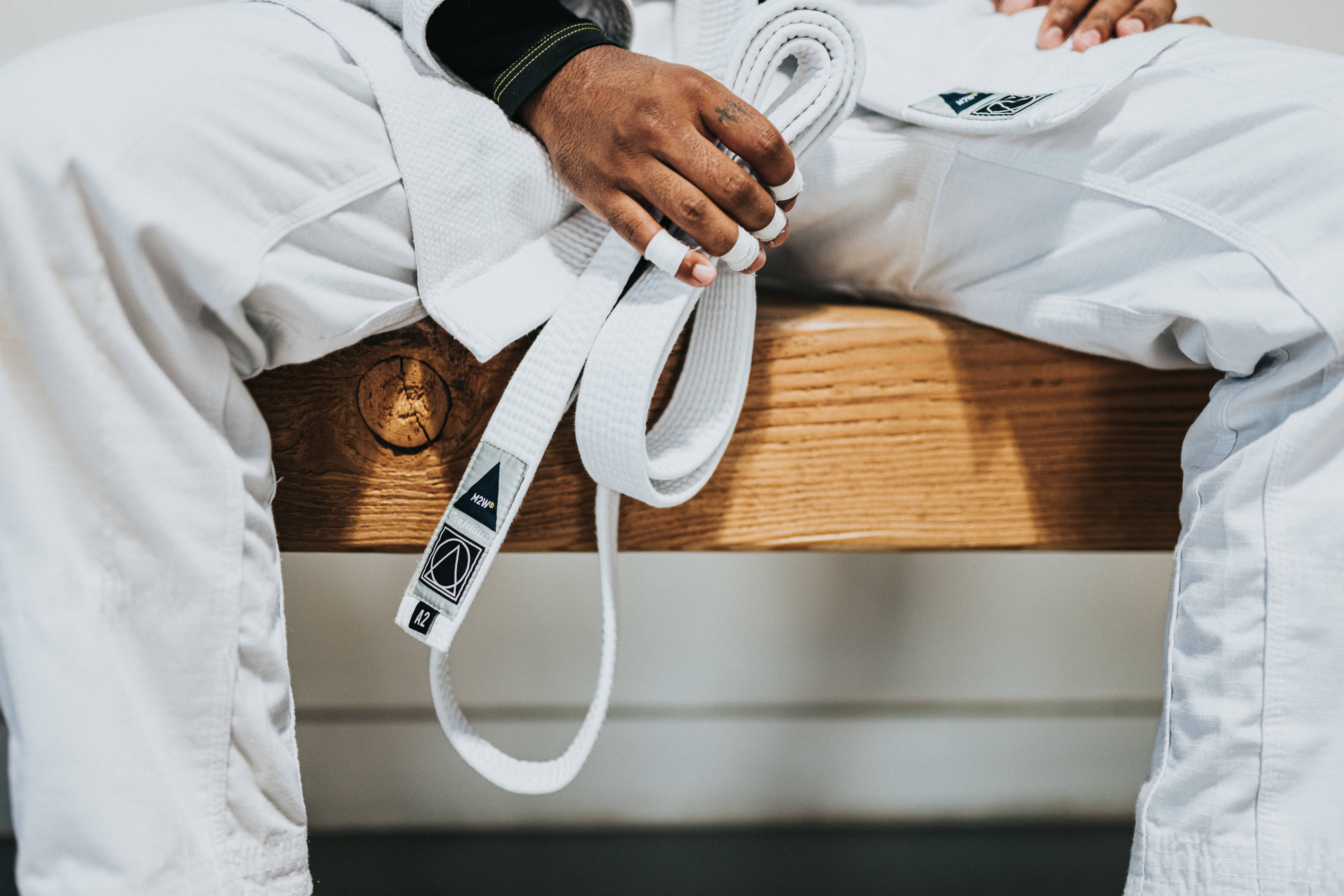 Judo 桌布 壁紙 — 一名身穿白色武術道服的人坐在木長凳上，手持帶有品牌標誌的白色腰帶。
