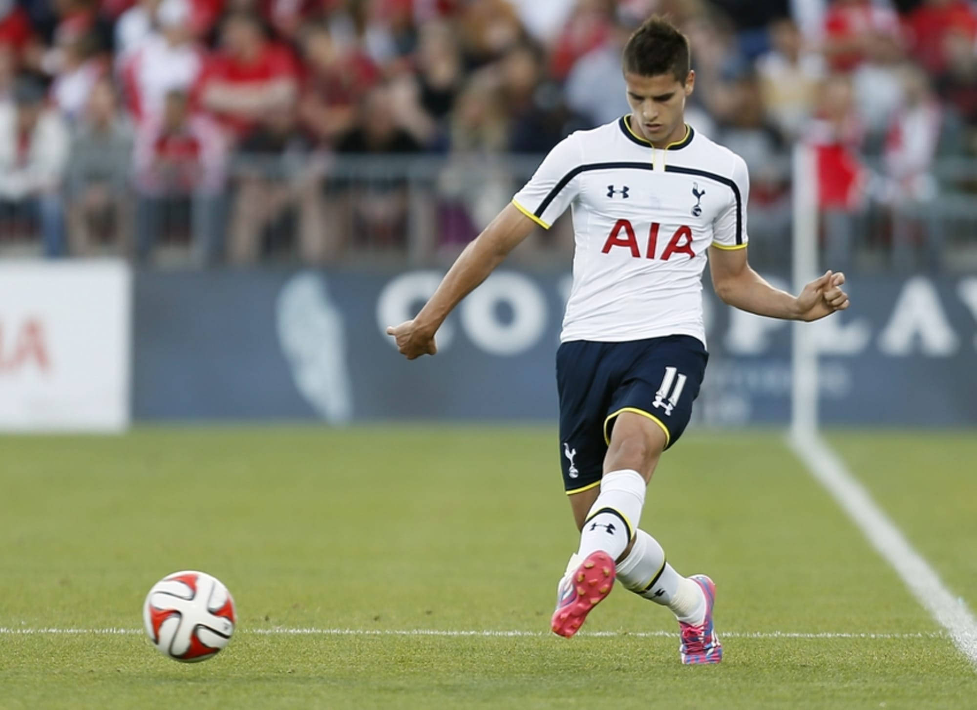 erik lamela 桌布 壁紙 — 一名身穿白色熱刺球衣的足球員準備在充滿活力的綠色球場上踢一個紅白相間的球，背景有模糊的觀眾。