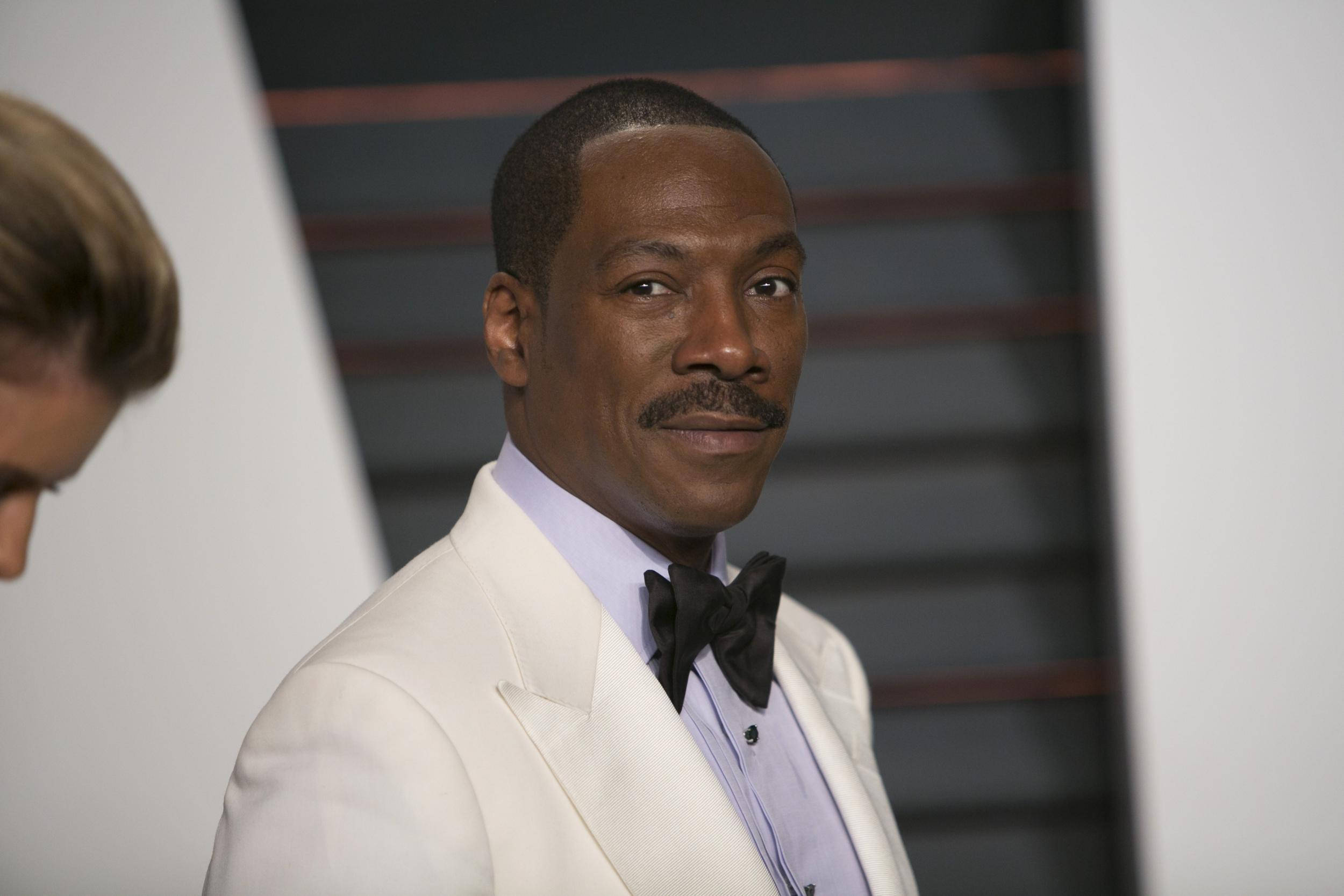 eddie murphy 桌布 壁紙 — 一名身穿白色燕尾服、繫著黑色領結的男子，在條紋背景前露出微妙微笑。