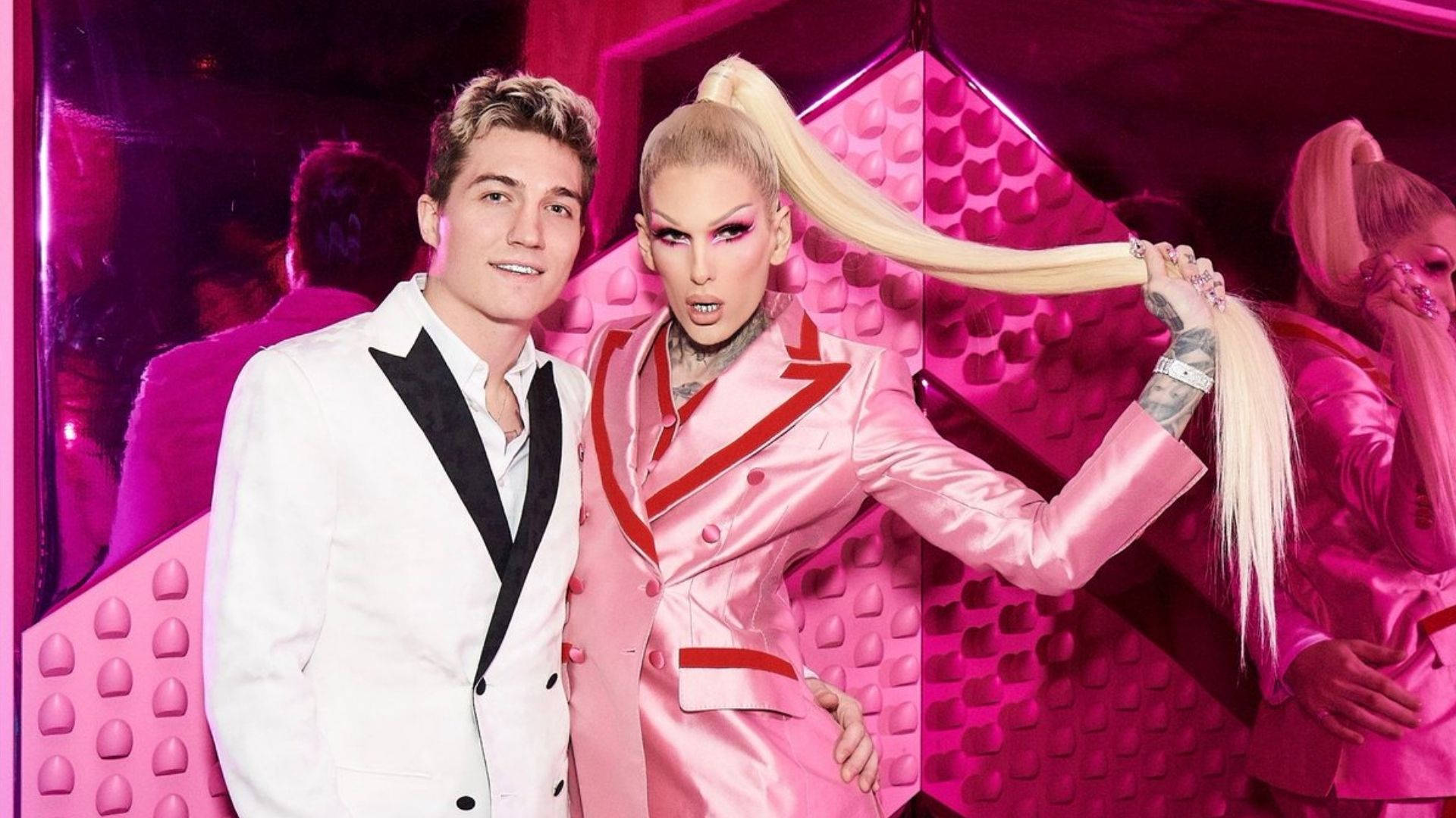 Jeffree Star 桌布 壁紙 — 一名身穿白色燕尾服的男子站在一位穿著粉紅色緞面西裝的變裝皇后旁邊，她將自己的金色長馬尾辮靠在粉紅色紋理牆上。