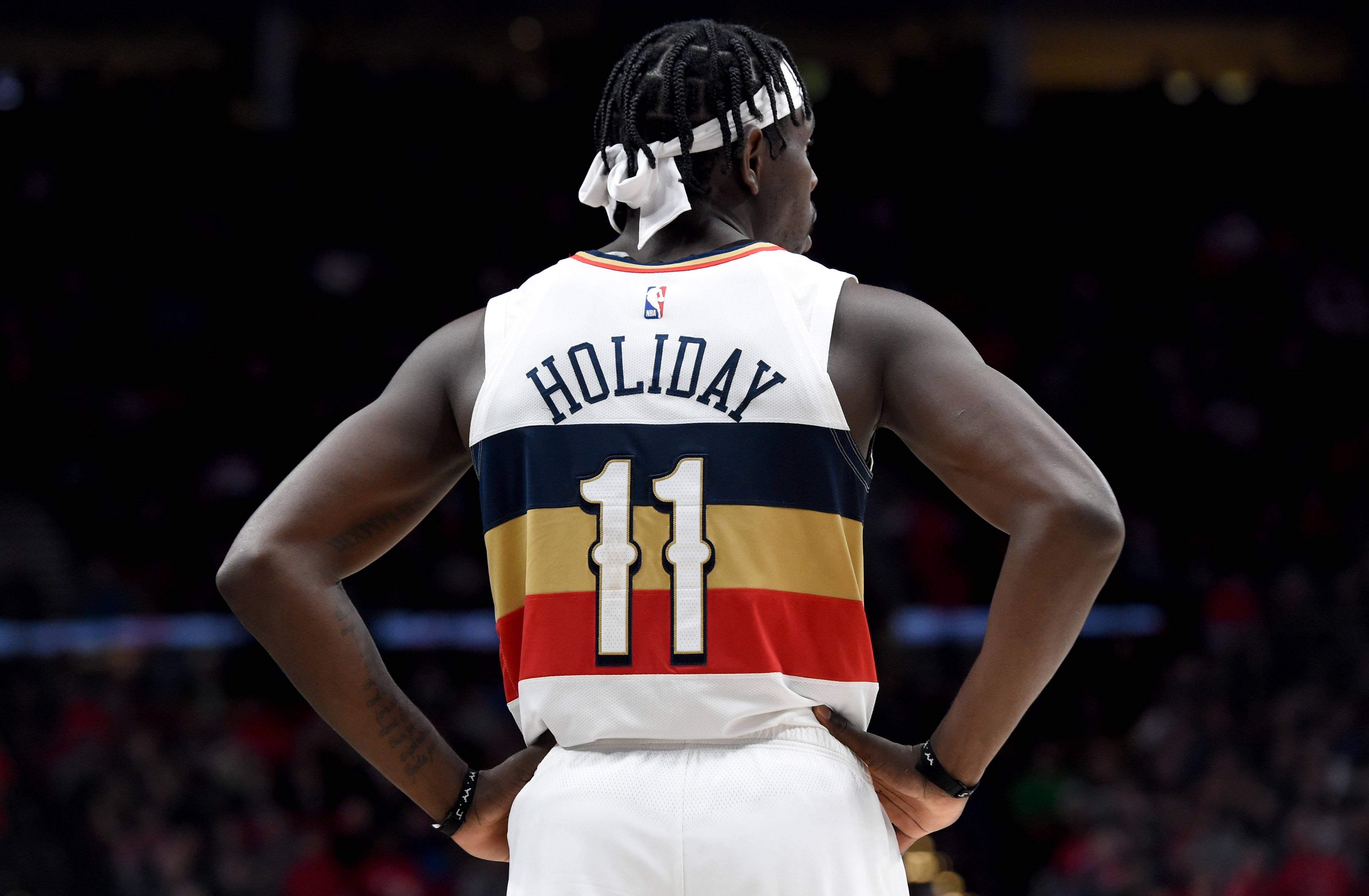 Jrue Holiday 桌布 壁紙 — 一名身穿白色球衣、印有「HOLIDAY 11」字樣的籃球運動員，在燈光昏暗的球場上背對鏡頭，雙手叉腰站立。