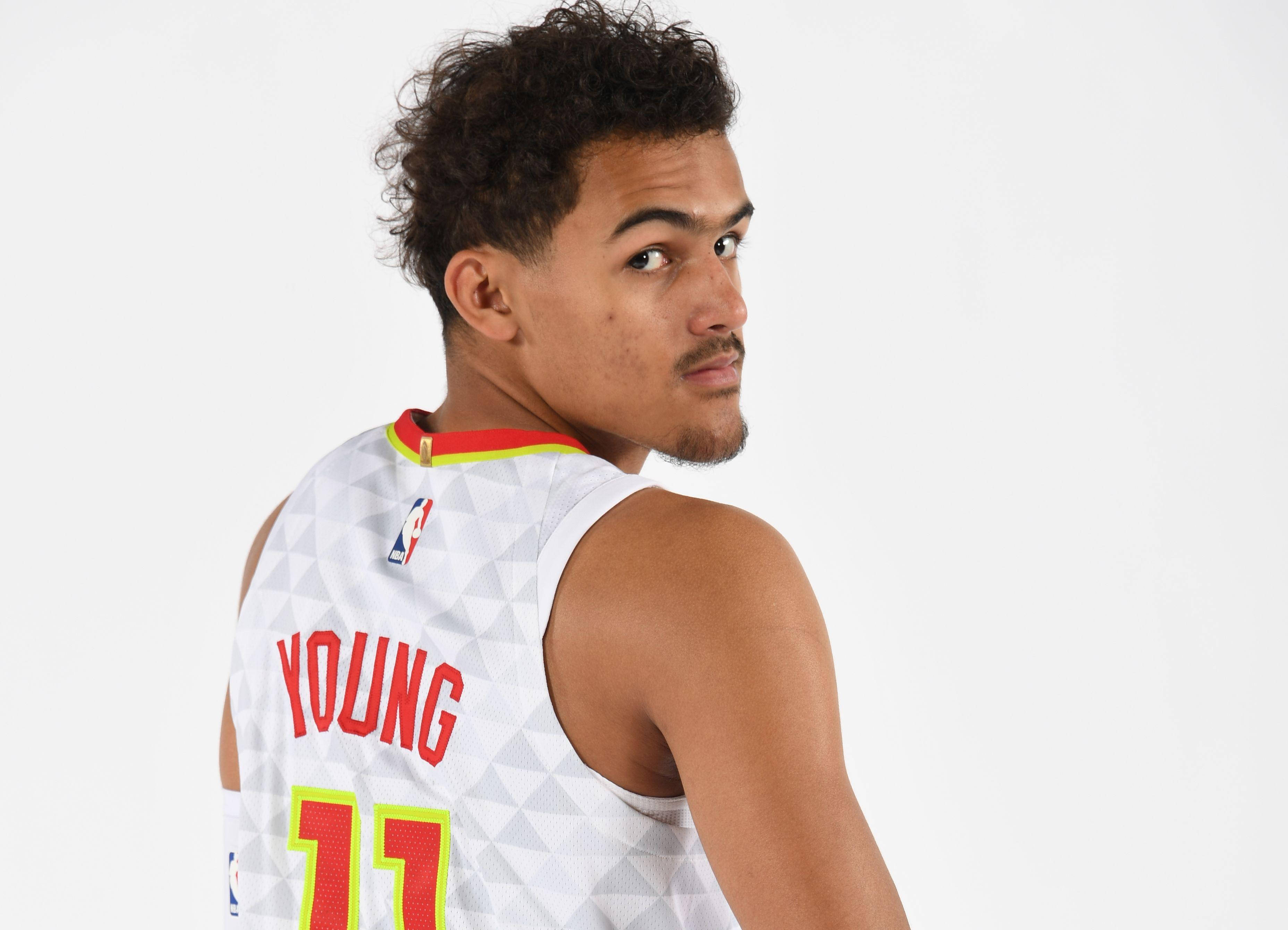 Trae Young 桌布 壁紙 — 一名身穿白色球衣、帶有紅色和黃色點綴的籃球運動員，在純白色背景上轉頭越過肩膀看向鏡頭。