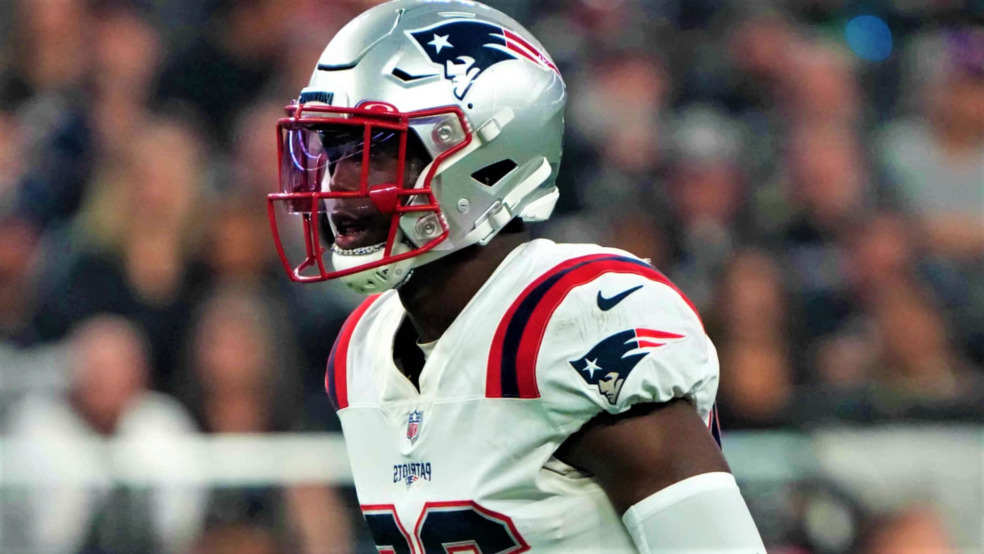 Devin Mccourty 桌布 壁紙 — 一名身穿白色球衣、戴著銀色頭盔和紅色面罩的新英格蘭愛國者隊美式足球員在夜間比賽中神情專注。