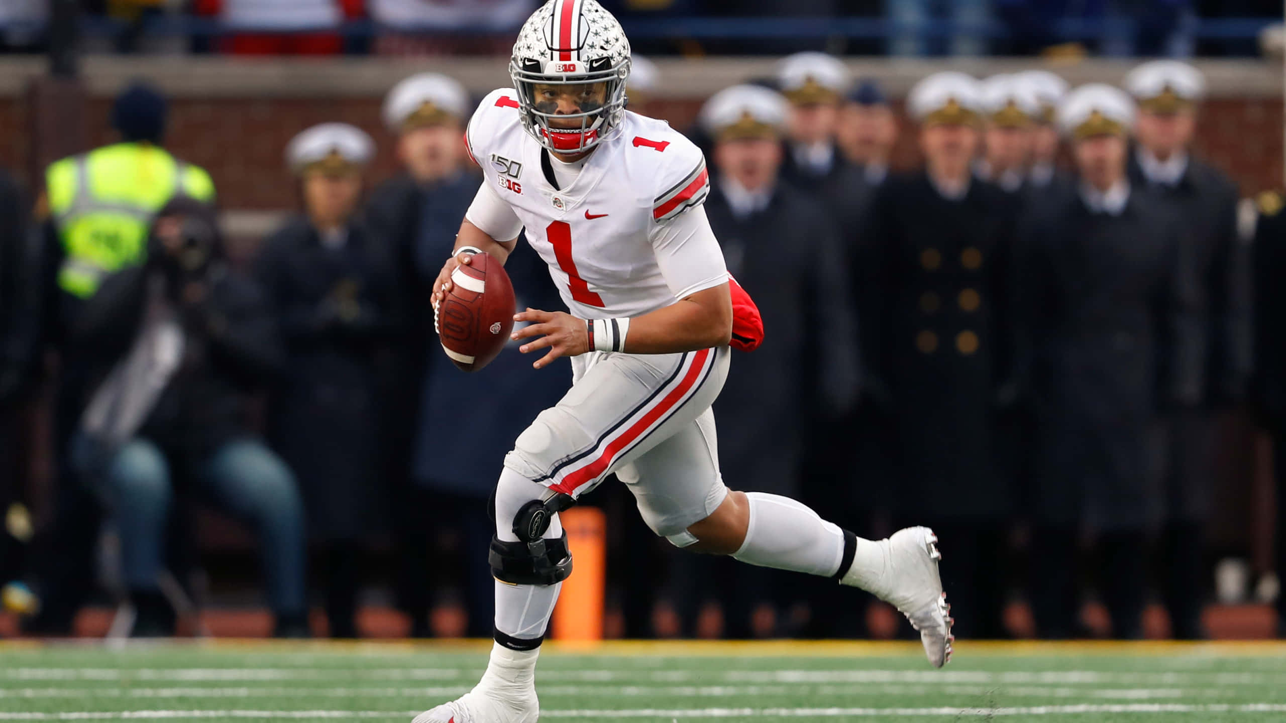 Justin Fields 桌布 壁紙 — 一名身穿白色球衣、背號1號的美式足球四分衛在綠色球場上帶球奔跑，背景有穿著制服的人員站立。