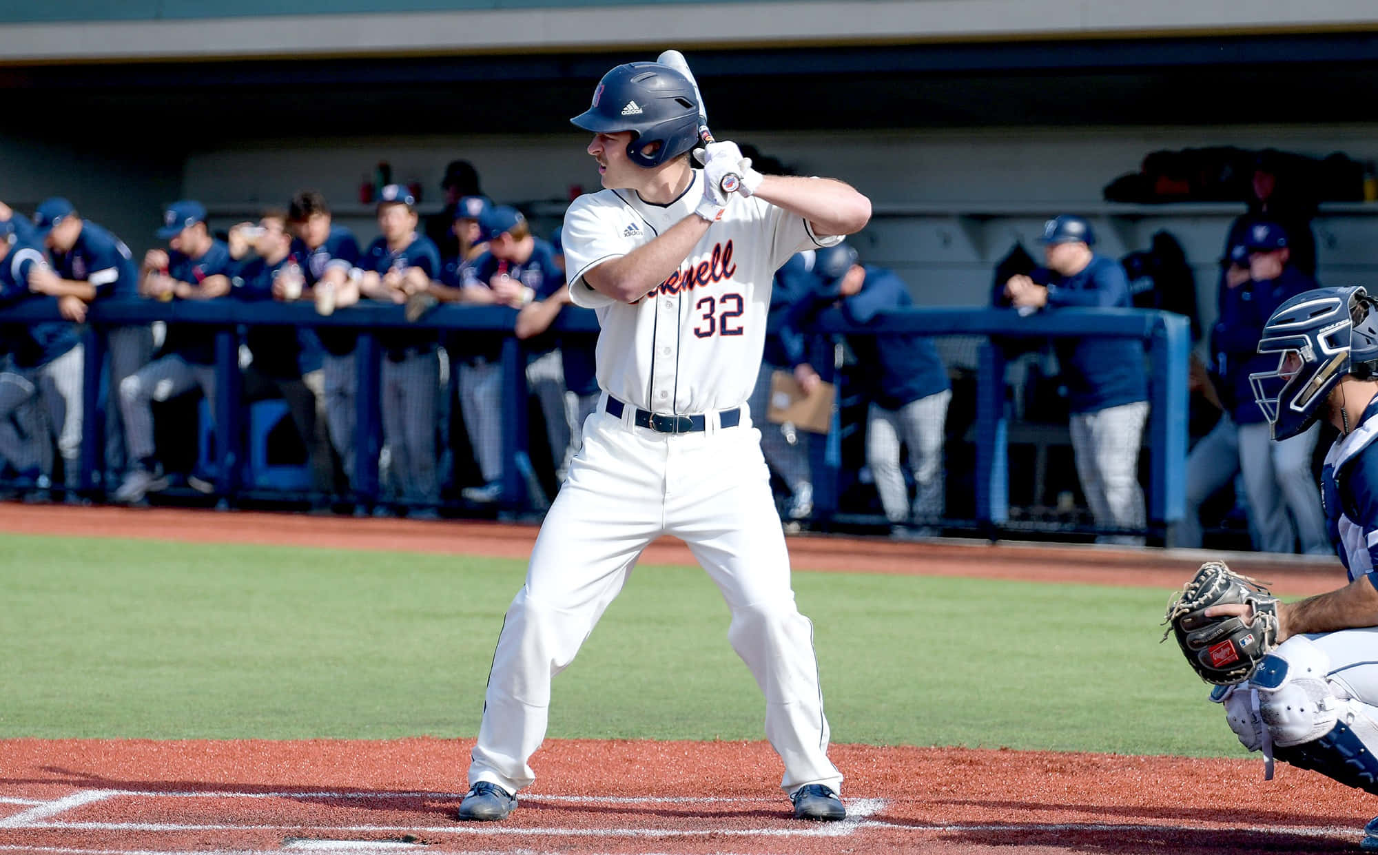 College Baseball 桌布 壁紙 — 一名身穿白色球衣、背號32的棒球選手在陽光明媚的日間比賽中揮棒，隊友在休息區觀看。