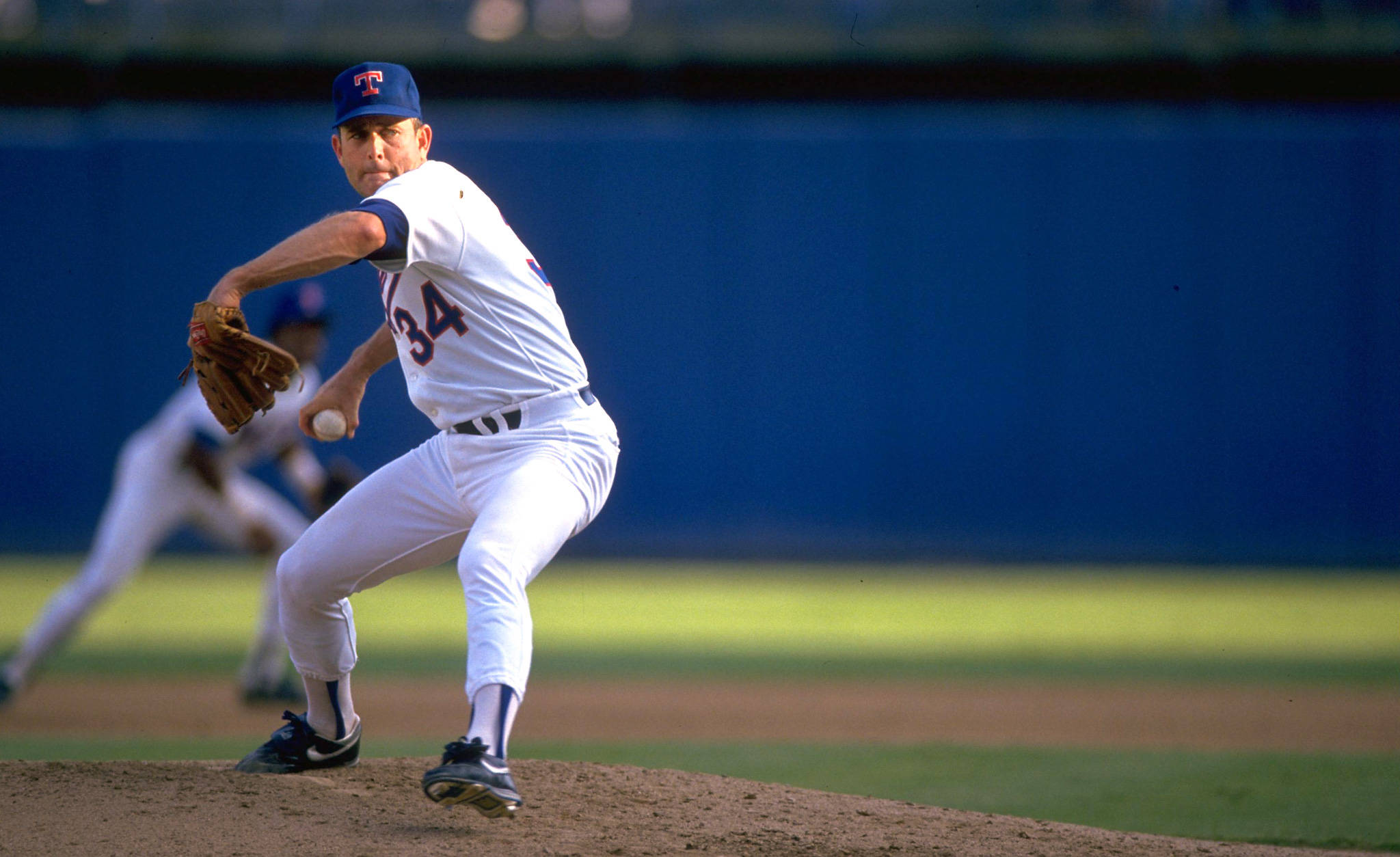 Nolan Ryan 桌布 壁紙 — 一名身穿白色球衣、背號34的棒球投手在投手丘上擺出投球姿勢，背景是藍色牆壁和綠色球場。