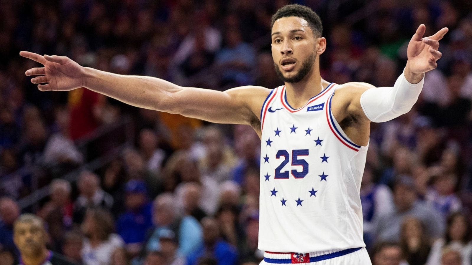 Ben Simmons 桌布 壁紙 — 一名身穿白色球衣、號碼25號、帶有藍色星星的籃球運動員在比賽中用力做手勢，背景是模糊的人群。