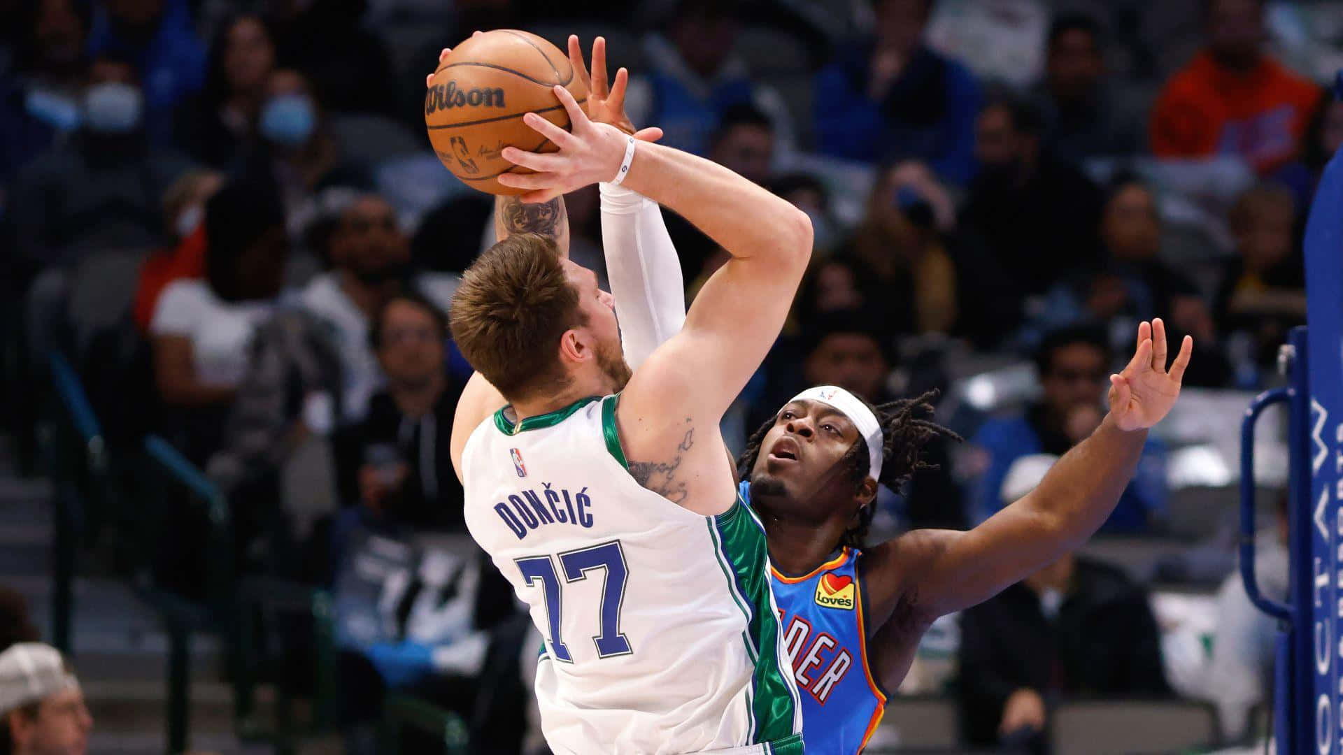 Luka Doncic 桌布 壁紙 — 一名身穿白色球衣、號碼77的籃球運動員正在投籃，同時被一名身穿藍橙色球衣的對手防守。