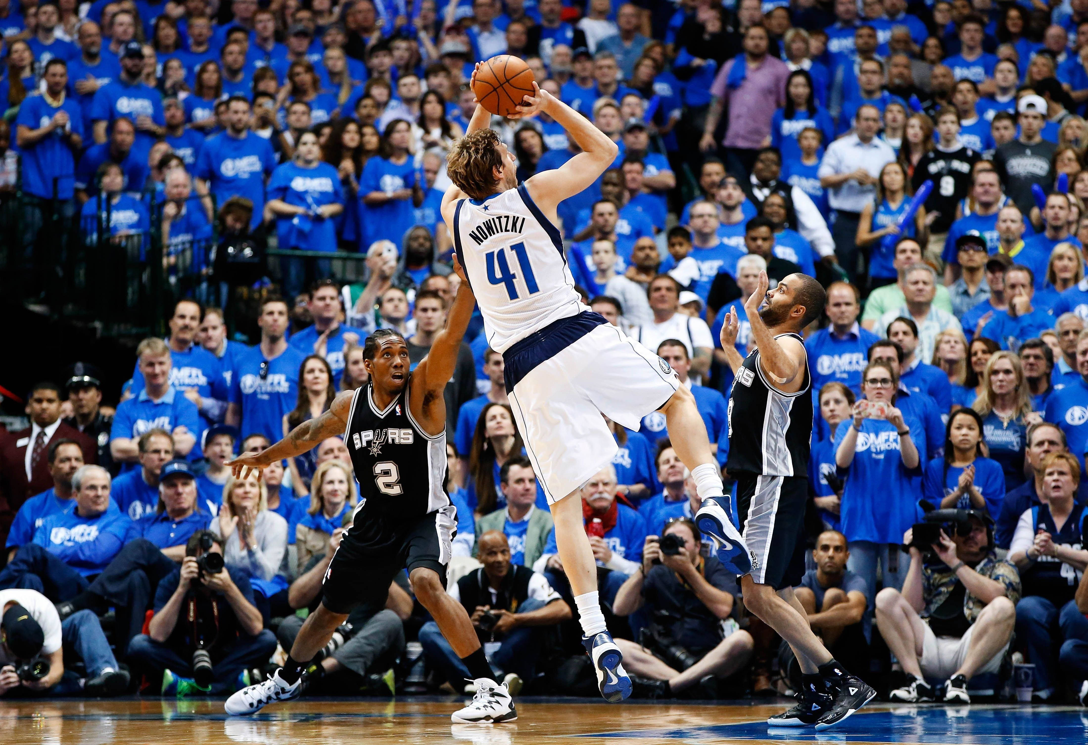 Dirk Nowitzki 桌布 壁紙 — 一名身穿白色球衣41號的籃球運動員高高躍起投籃，身穿黑色球衣的防守球員試圖封阻他，背景是一群身穿藍色衣服的球迷。