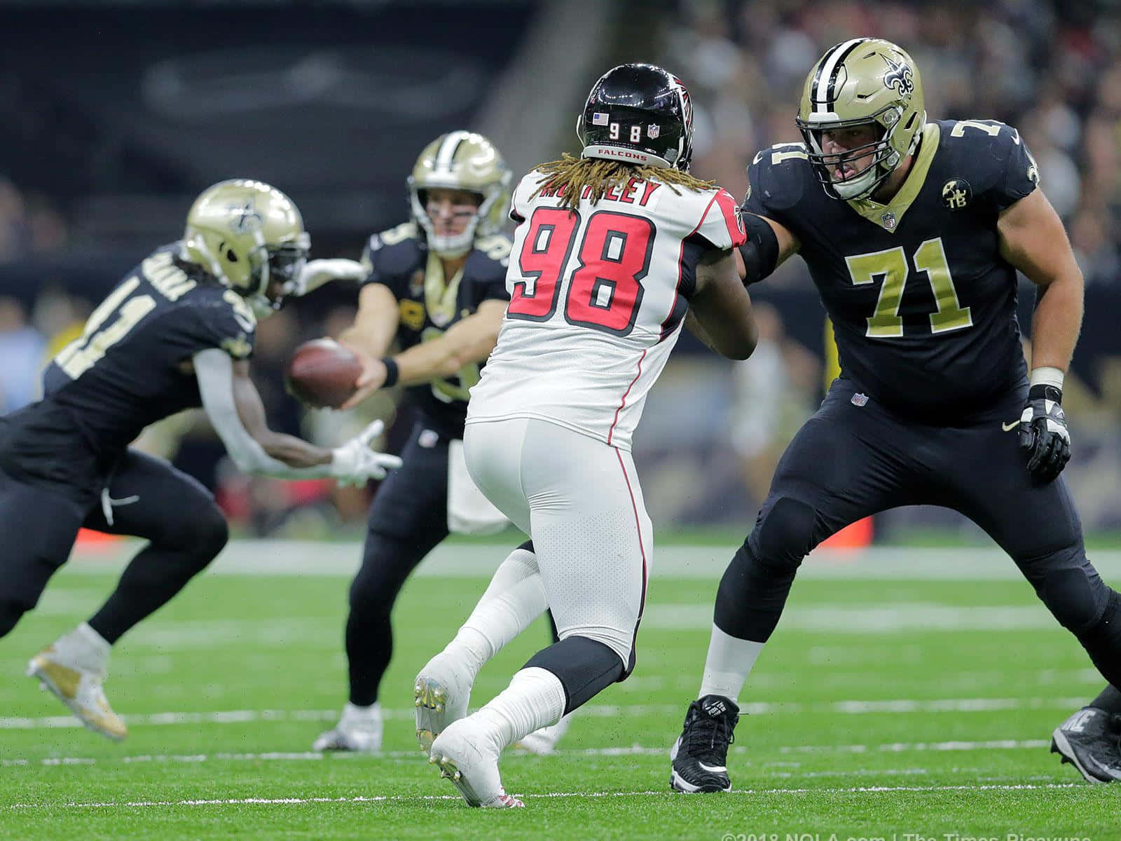 Atlanta Falcons 桌布 壁紙 — 一名身穿白色球衣98號的美式足球員阻擋一名黑色球衣的對手，而四分衛正在準備在球場燈光下投球。