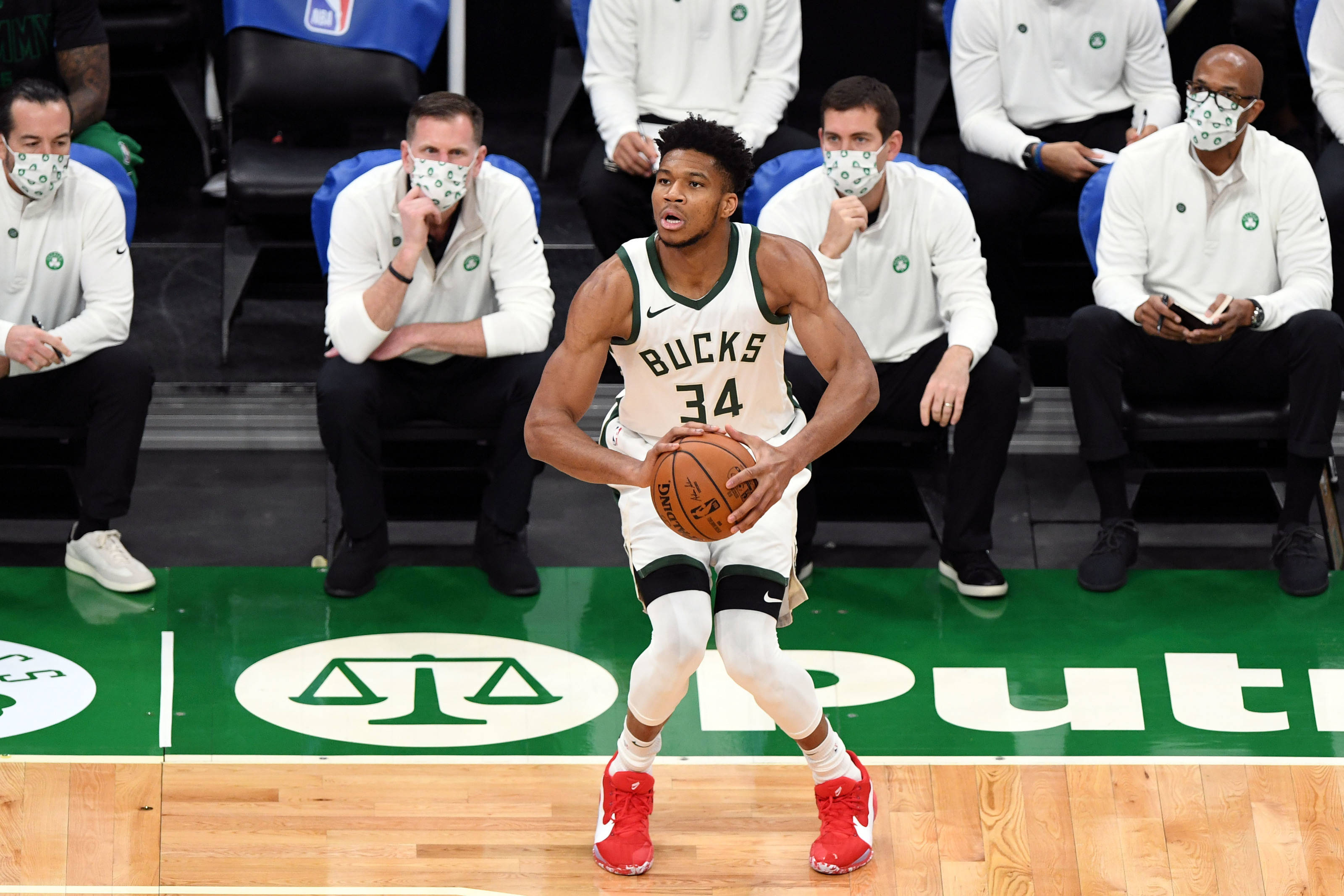 Giannis 桌布 壁紙 — 一名身穿白色球衣和紅色球鞋的密爾瓦基公鹿籃球球員在綠色球場上運球，戴著口罩的教練在板凳席觀看。