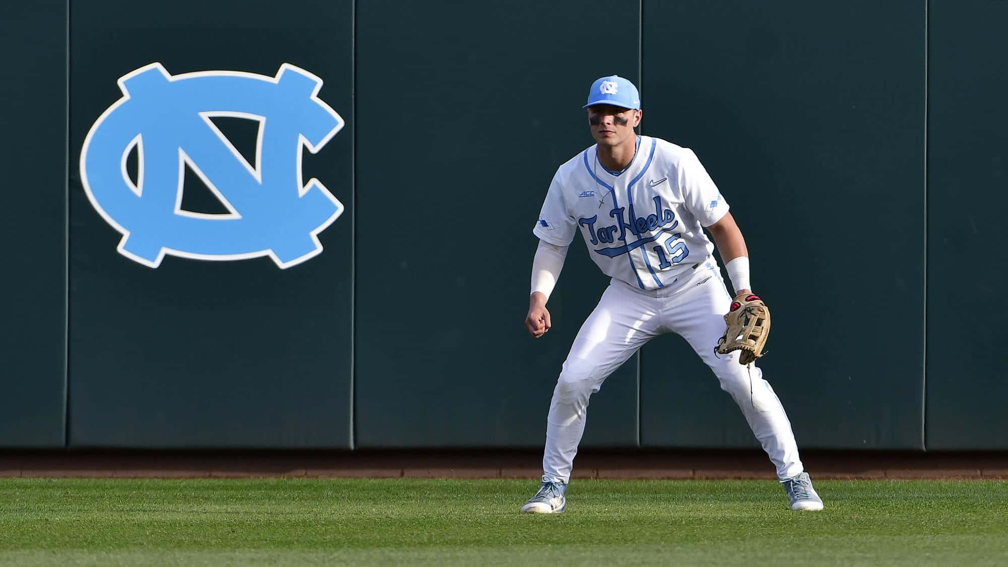 College Baseball 桌布 壁紙 — 一名身穿白色球衣和藍色帽子的棒球運動員警覺地站在綠色草地上，靠近一面帶有大型淺藍色NC標誌的深色牆壁。