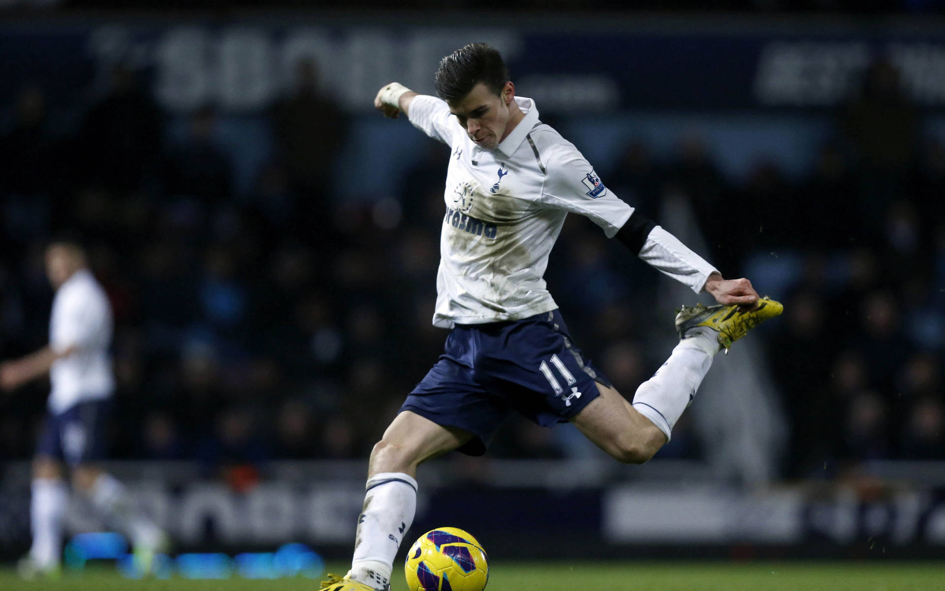 Gareth Bale 桌布 壁紙 — 一名身穿白色球衣和藍色短褲的足球運動員在夜間比賽中，在體育場燈光下凌空踢起一個黃紫相間的球。