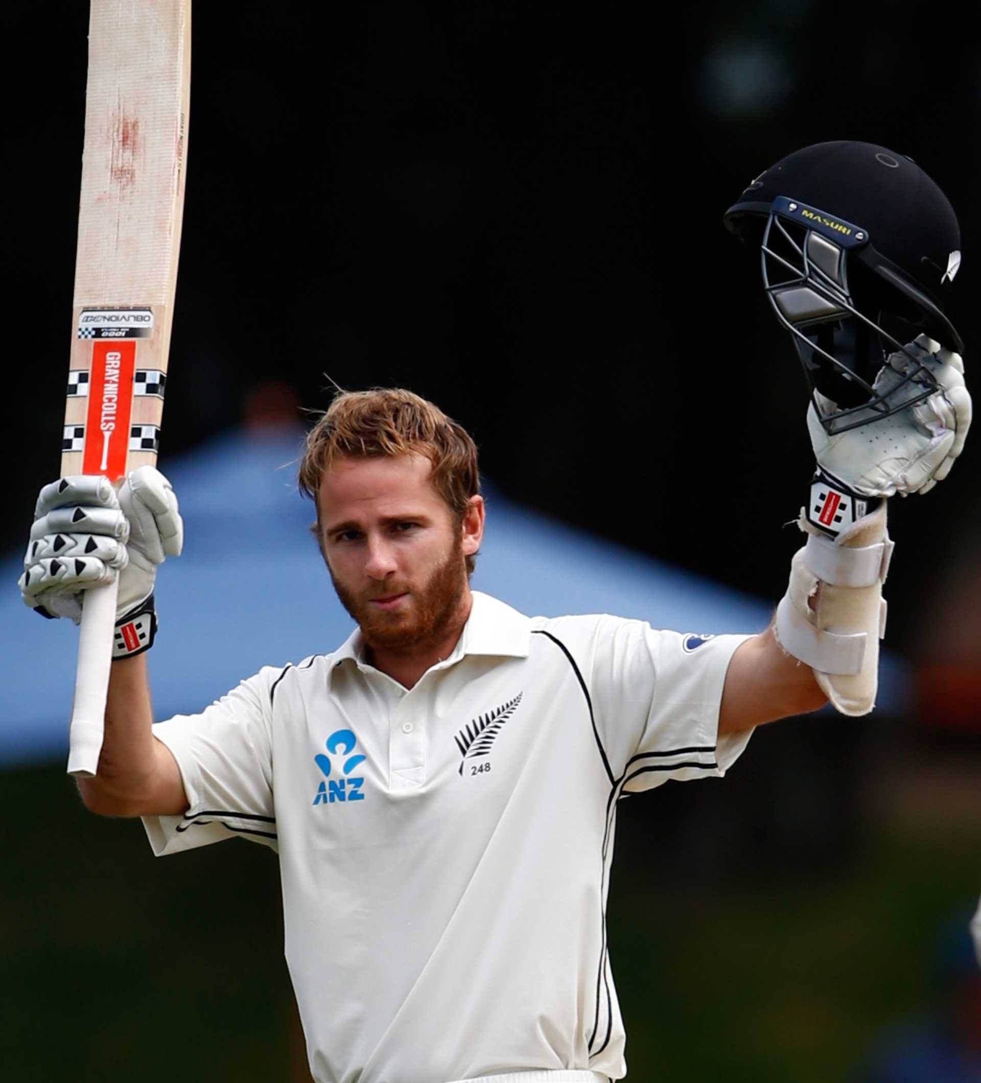 kane williamson 桌布 壁紙 — 一名身穿白色球衣的板球運動員在模糊的體育場背景下，高舉球棒和頭盔慶祝勝利。