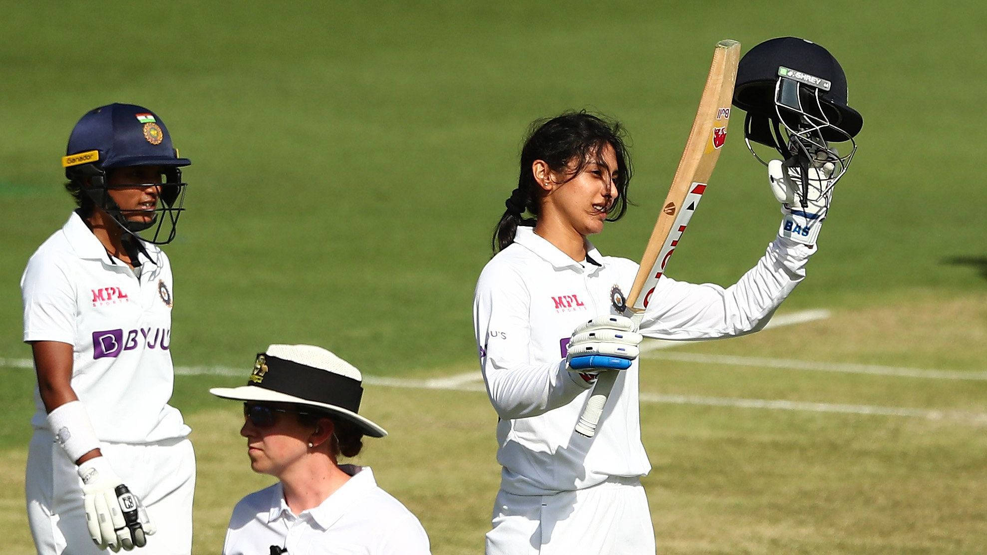 Smriti Mandhana 桌布 壁紙 — 一名身穿白色球衣的板球運動員在陽光明媚的綠色球場上高舉球棒和頭盔慶祝勝利，隊友和裁判在附近。