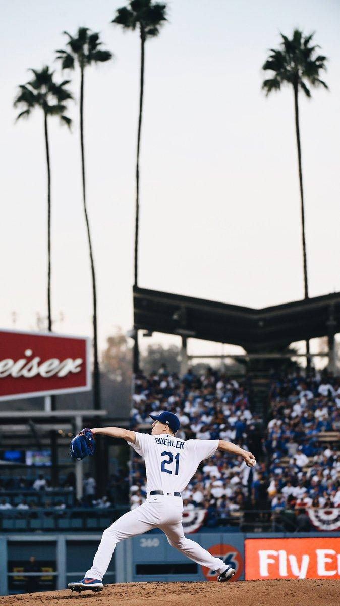 walker buehler 桌布 壁紙 — 一名身穿白色球衣的棒球投手在陽光明媚的投手丘上投球，身後有棕櫚樹和歡呼的人群。
