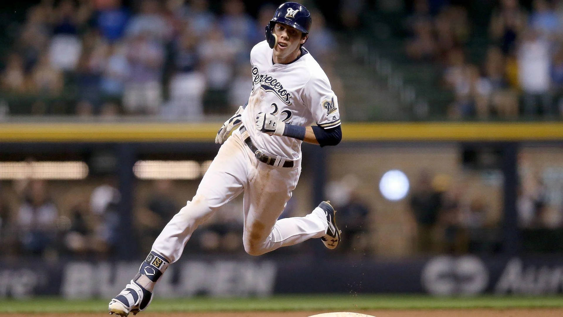Yelich 桌布 壁紙 — 一名身穿白色球衣的棒球運動員在球場上跨步跳躍，背景是模糊的體育場燈光和觀眾。