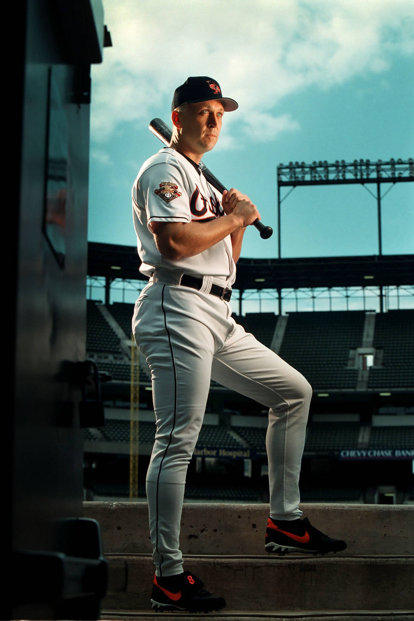 cal ripken jr 桌布 壁紙 — 一名身穿白色球衣的棒球運動員自信地站在體育場的球員休息區，手持球棒，背景是明亮的藍天。