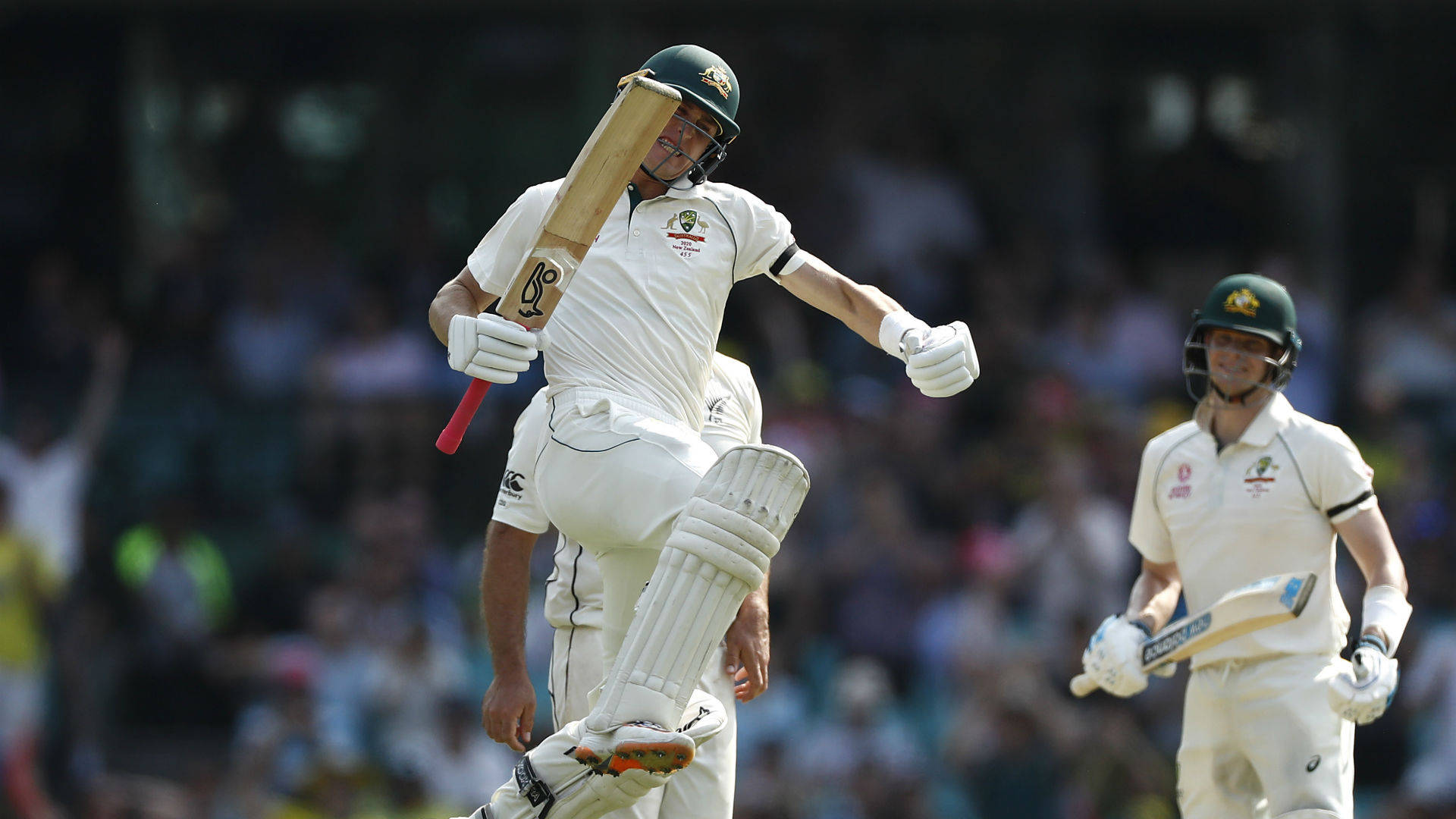 australia cricket 桌布 壁紙 — 一名身穿白色球衣的澳洲板球運動員在歡呼的人群中高舉球棒並抬起一條腿慶祝一個里程碑。