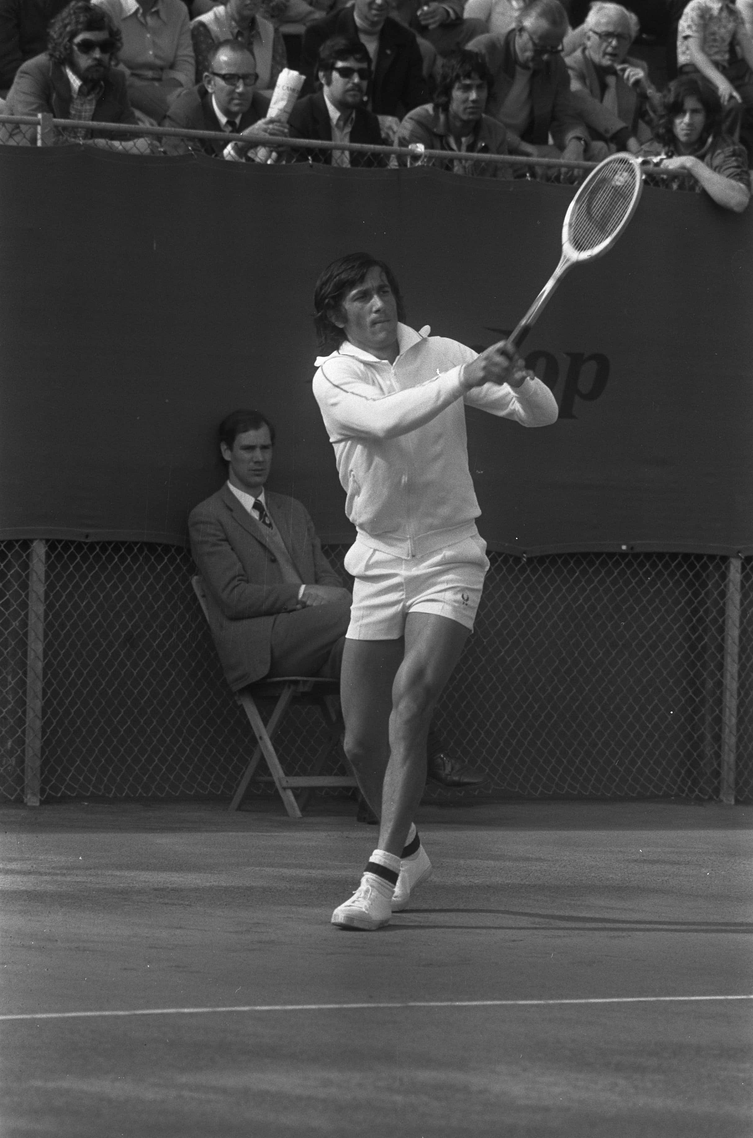 Tennis 桌布 壁紙 — 一名身穿白色球衣的男網球運動員在硬地球場上比賽中揮拍，觀眾在圍欄後觀看。