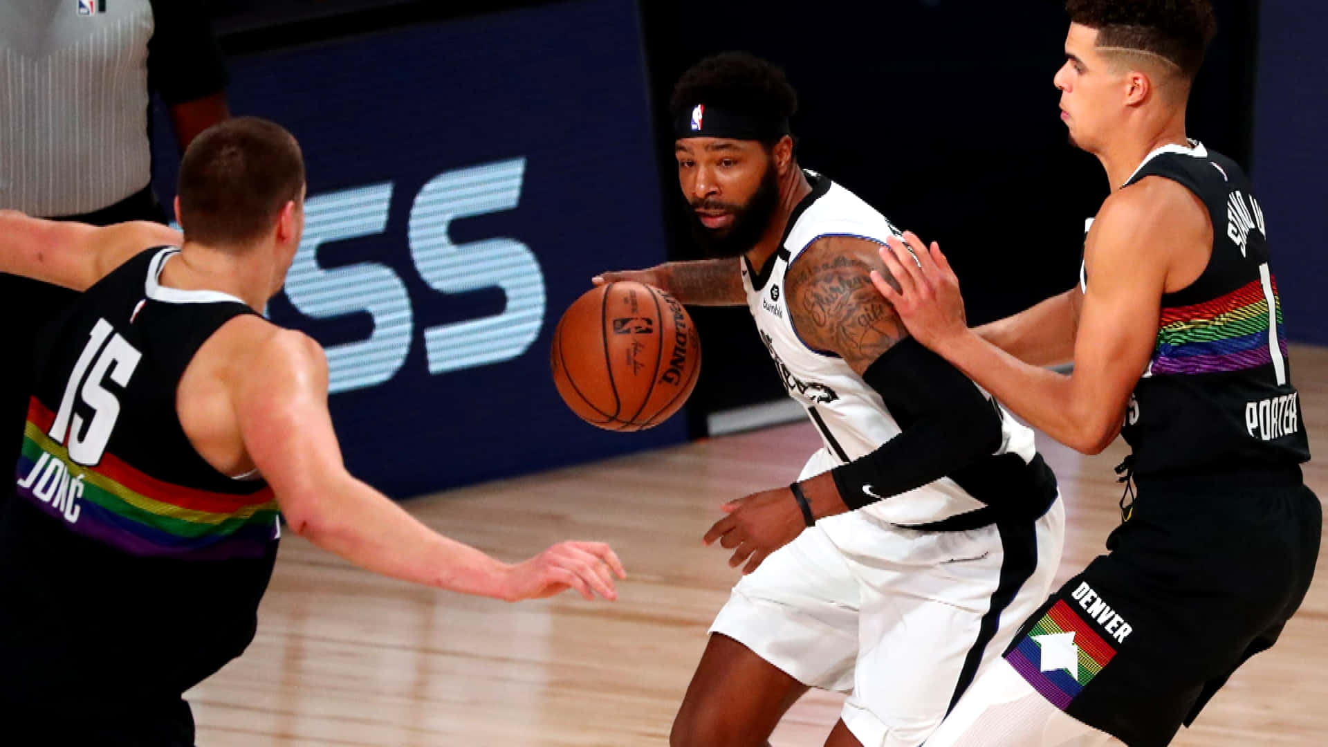 NBA 桌布 壁紙 — 一名身穿白色球衣的籃球運動員在光亮的球場上運球，同時被兩名身穿帶有彩虹色調黑色球衣的對手緊密防守。