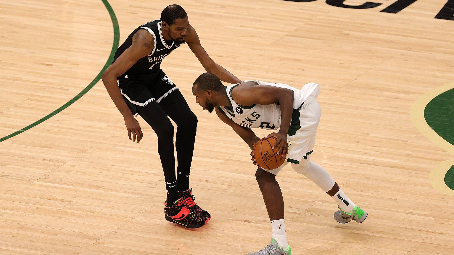 Khris Middleton 桌布 壁紙 — 一名身穿白色球衣的籃球運動員在拋光木地板上運球越過一名身穿黑色球衣的防守球員。