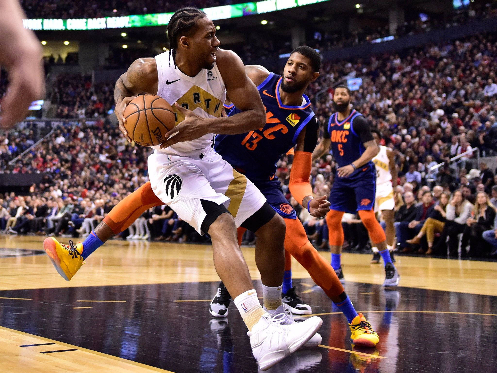 Kawhi 桌布 壁紙 — 一名身穿白色球衣的籃球運動員在擠滿觀眾的球場上，於激烈比賽中運球越過藍色球衣的防守球員。