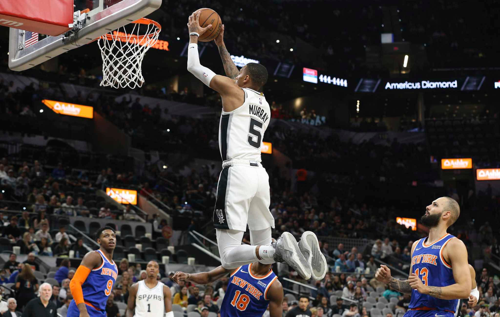 Dejounte Murray 桌布 壁紙 — 一名身穿白色球衣的籃球運動員在擠滿觀眾的場館中灌籃，防守球員從下方觀看。