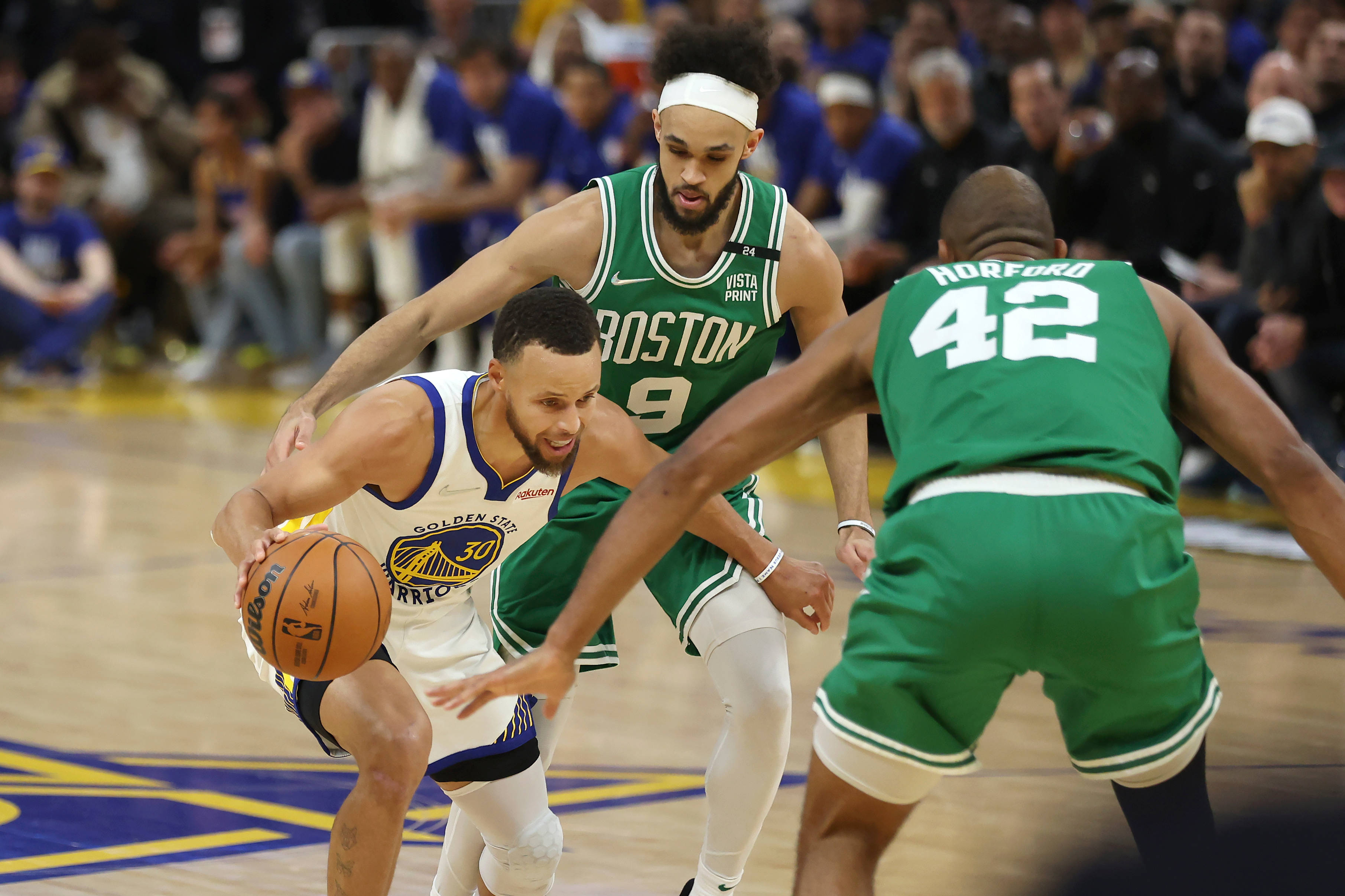 NBA 桌布 壁紙 — 一名身穿白色球衣的籃球運動員在激烈的比賽中運球，同時被兩名身穿綠色球衣的對手緊密防守。