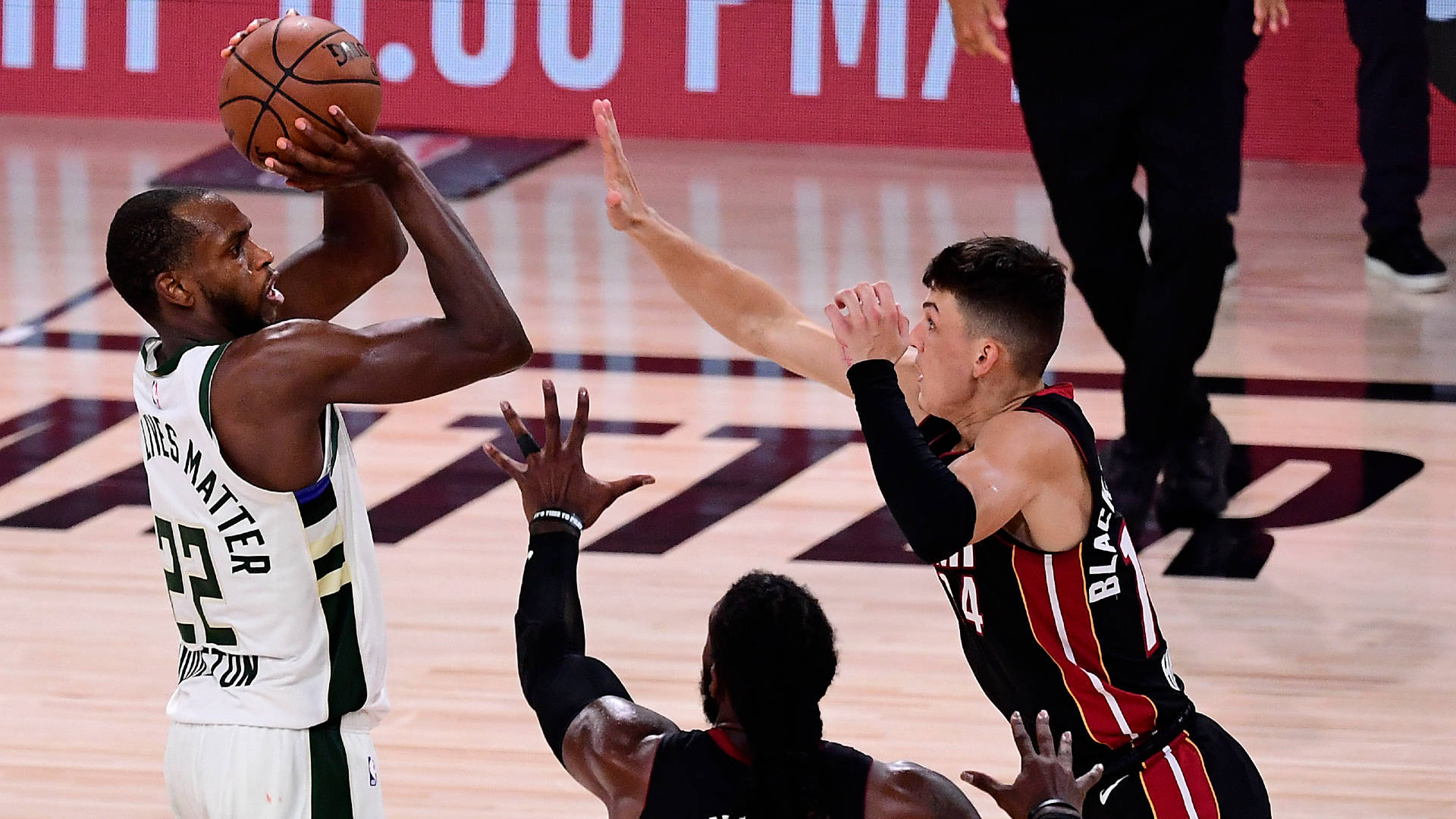 Khris Middleton 桌布 壁紙 — 一名身穿白色球衣的籃球運動員在燈光明亮的球場上，在對手的防守下投籃。