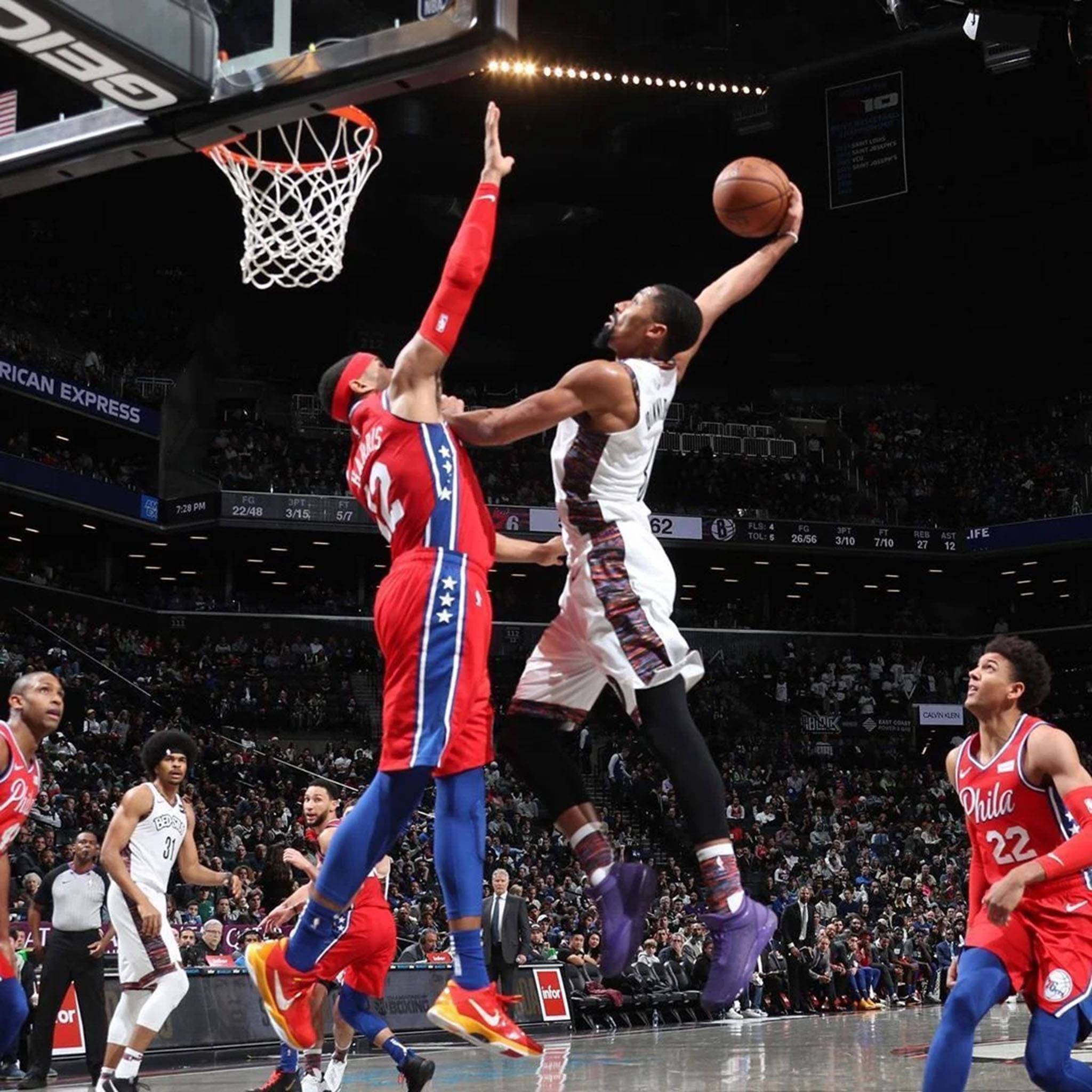 spencer dinwiddie 桌布 壁紙 — 一名身穿白色球衣的籃球運動員跳向籃框，而一名身穿紅色球衣的防守球員在擠滿觀眾的場館比賽中試圖封阻他的投籃。