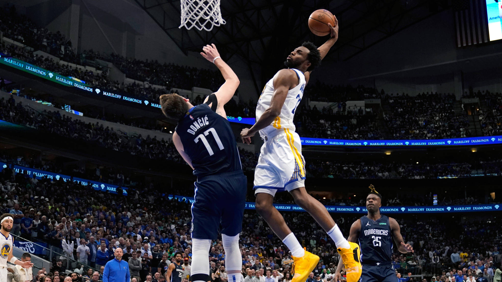 Andrew Wiggins 桌布 壁紙 — 一名身穿白色球衣的籃球運動員高高躍起準備灌籃，而一名身穿藍色球衣的防守球員在明亮的場館燈光下試圖封阻他。