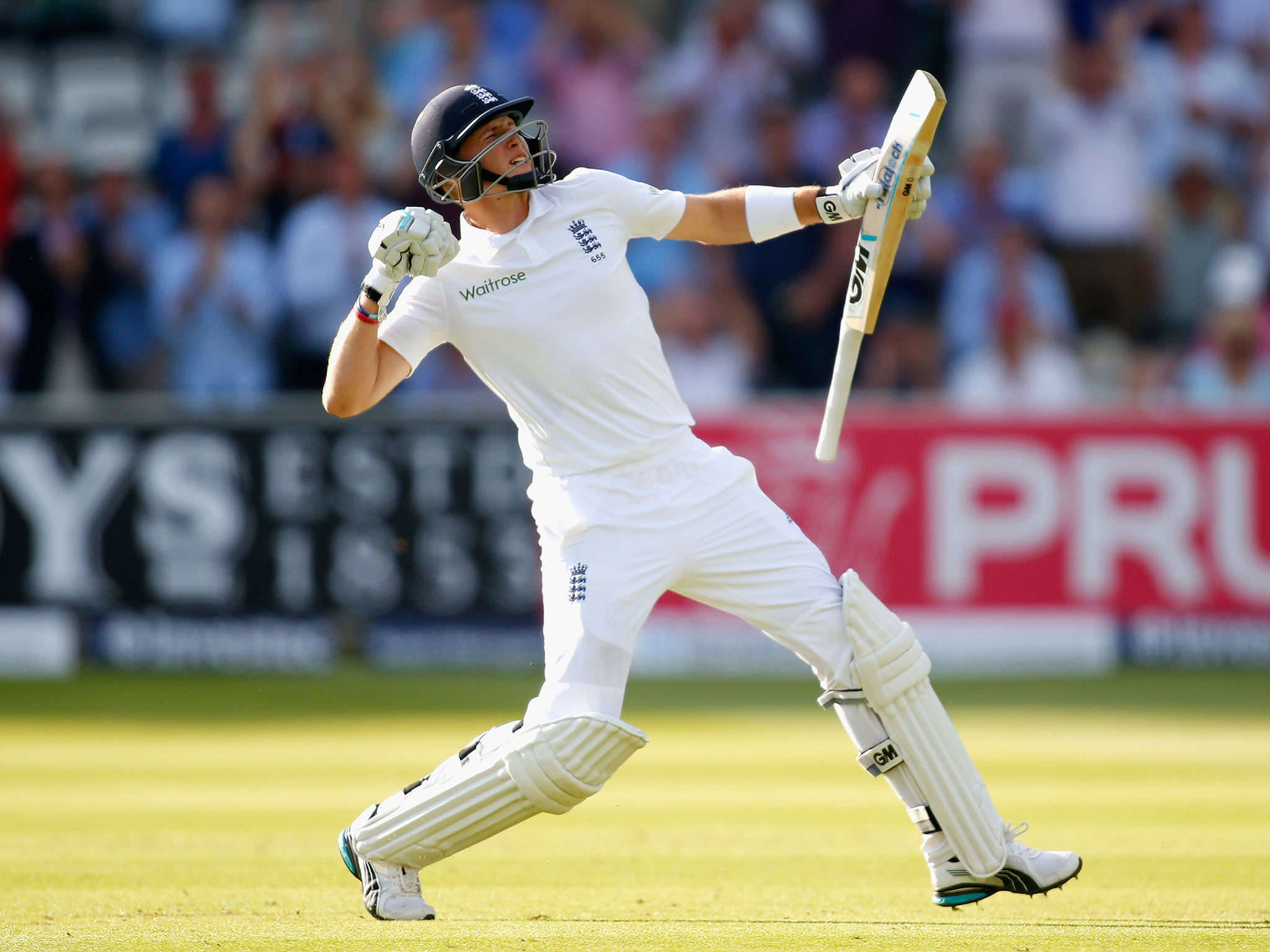England Cricket 桌布 壁紙 — 一名身穿白色球衣的英格蘭板球擊球手在陽光明媚的綠色球場上慶祝強力擊球，背景是模糊的觀眾。