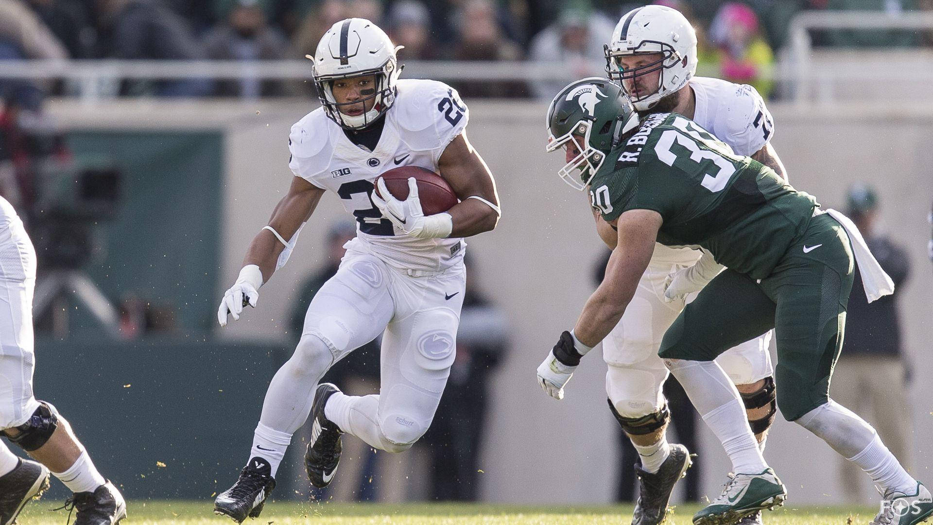 Saquon Barkley 桌布 壁紙 — 一名身穿白色球衣的足球員帶著球奔跑，綠色防守球員在陽光明媚的球場上飛身鏟球。
