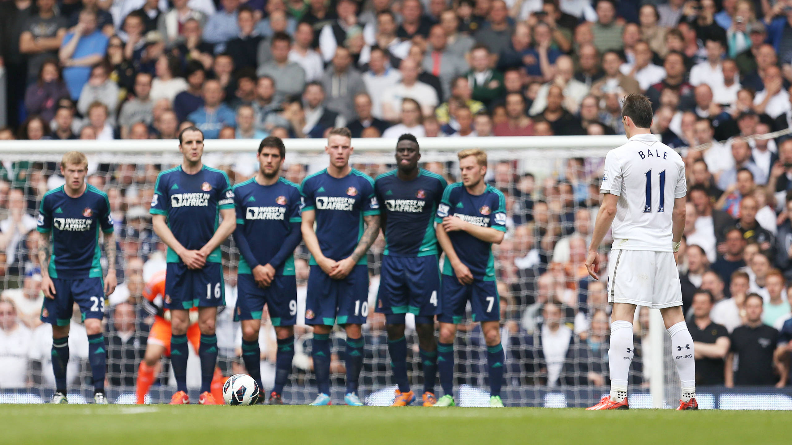 Gareth Bale 桌布 壁紙 — 一名身穿白色球衣的足球員獨自站在陽光普照的球場上，面對一排身穿藍色和藍綠色球衣的對方球員。