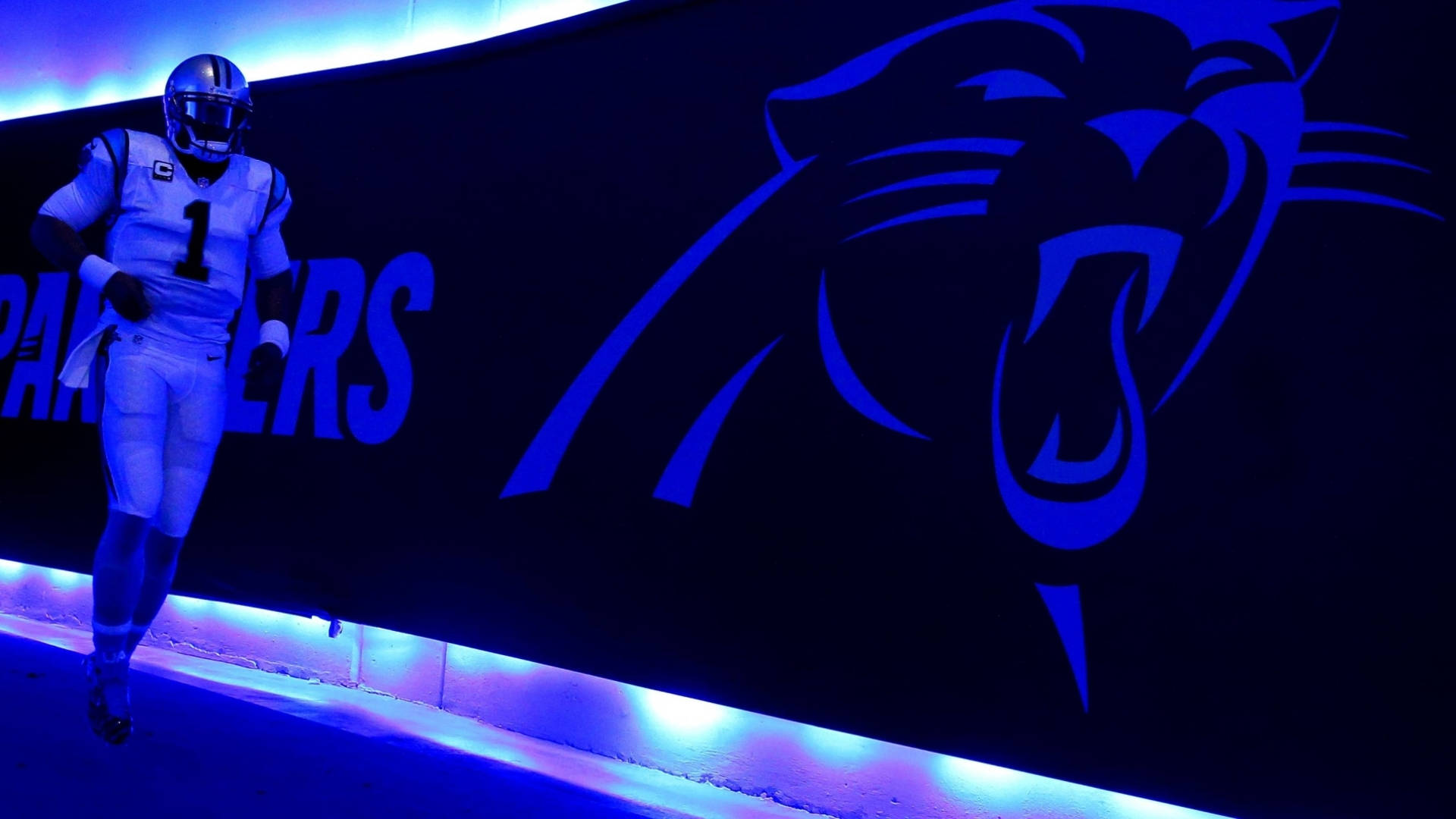 Panthers 桌布 壁紙 — 一名身穿白色球衣的足球運動員在黑暗隧道牆上發光的藍色黑豹標誌旁跑過。