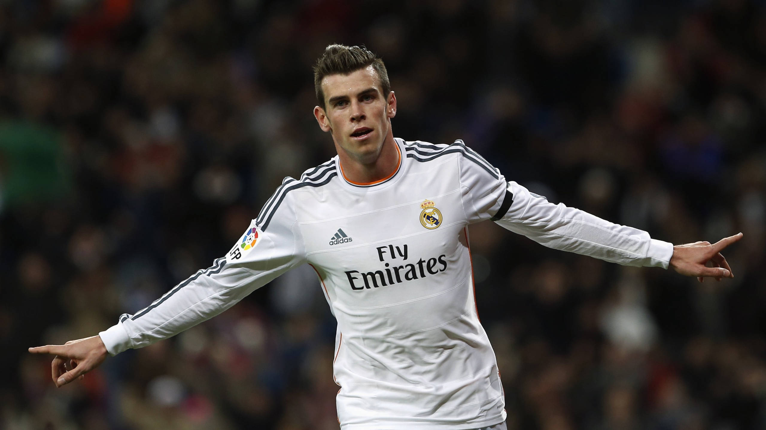 Gareth Bale 桌布 壁紙 — 一名身穿白色皇家馬德里球衣的足球員在模糊的球場人群背景下張開雙臂慶祝。