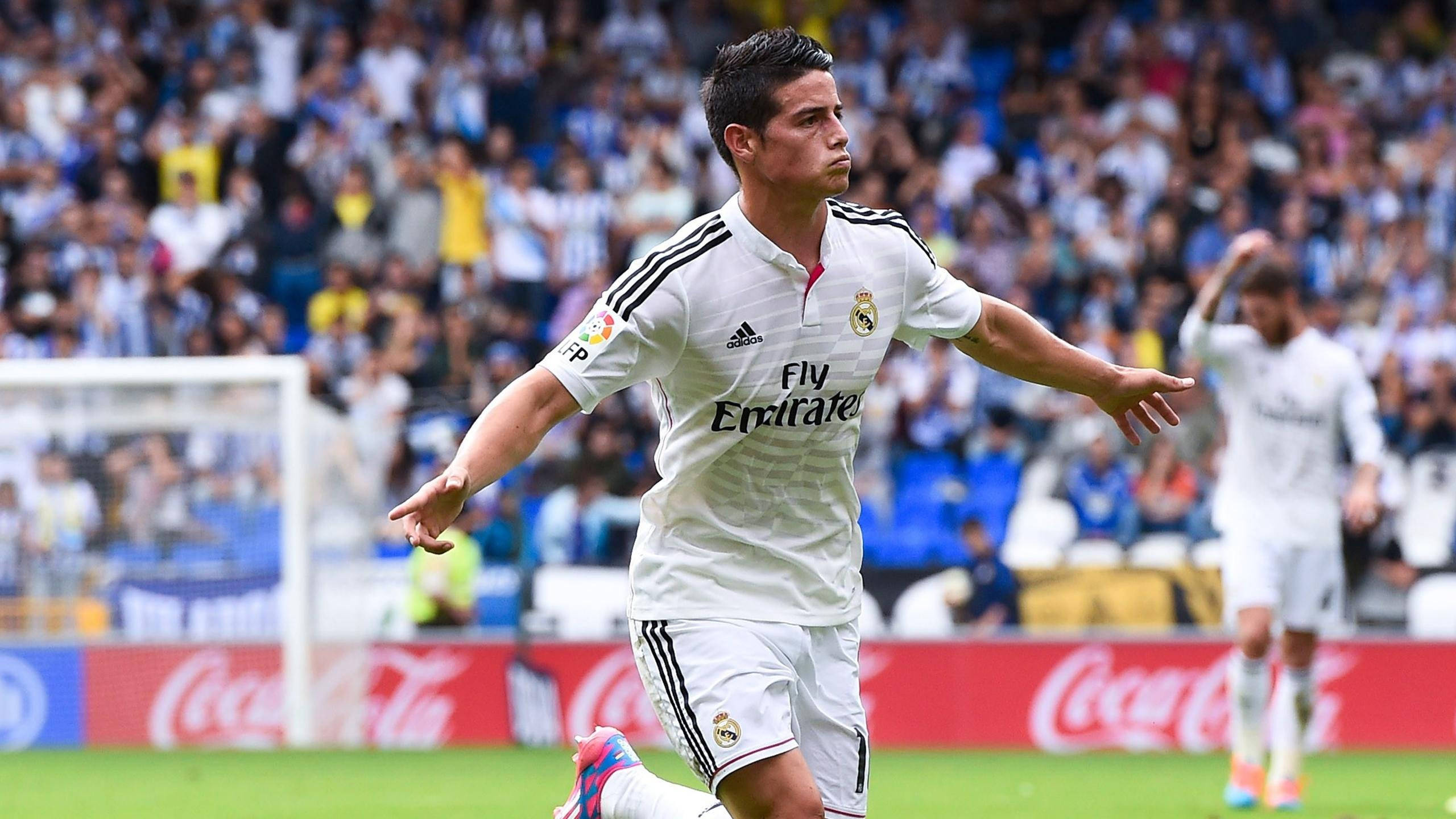 James Rodriguez 桌布 壁紙 — 一名身穿白色皇家馬德里球衣的足球運動員在陽光明媚的綠色球場上慶祝進球，身後是座無虛席的體育場。