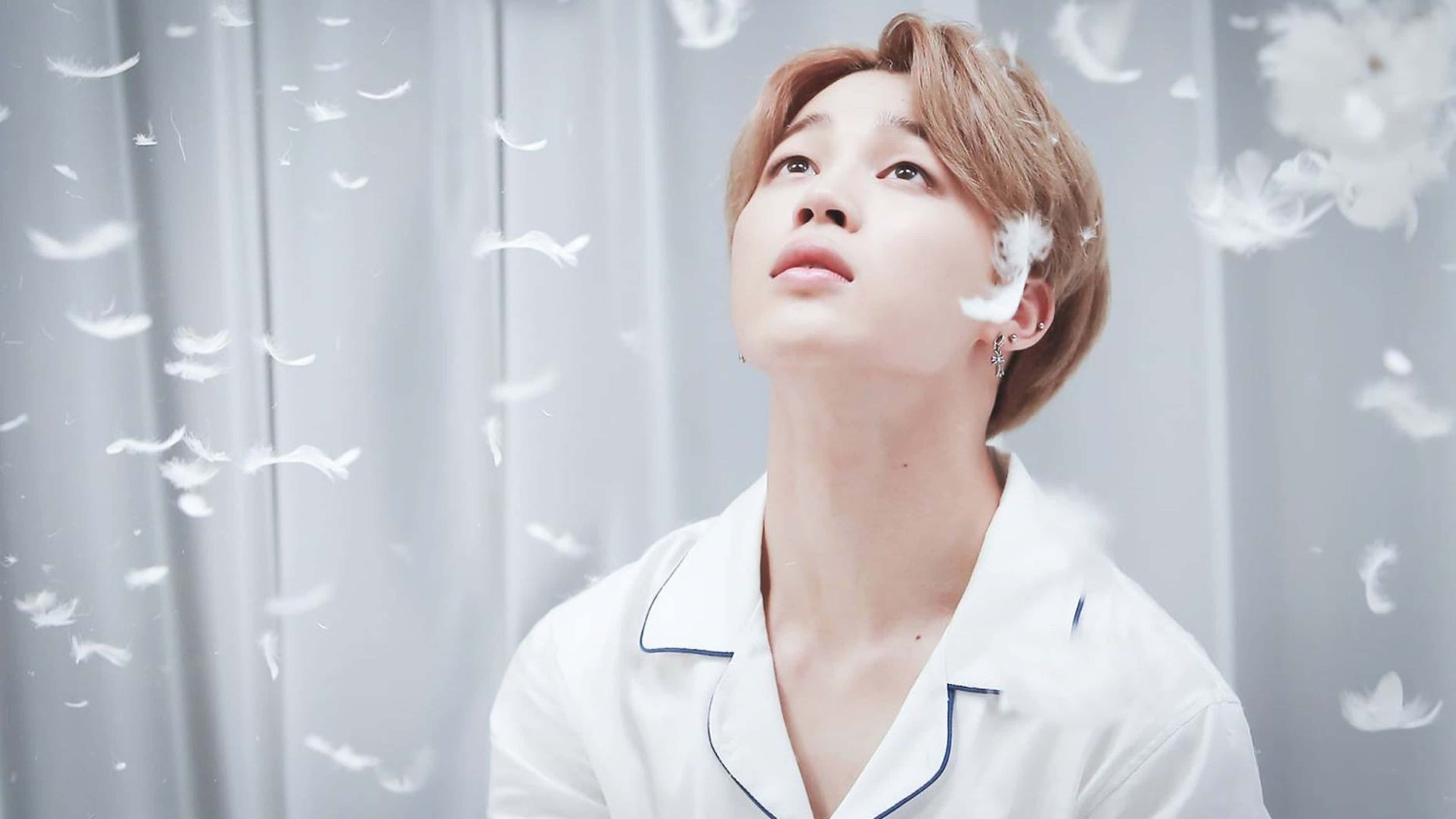 Bts Jimin 桌布 壁紙 — 一名身穿白色睡衣的年輕男子向上凝視，柔軟的白色羽毛在他周圍飄浮，背景是淺灰色窗簾。