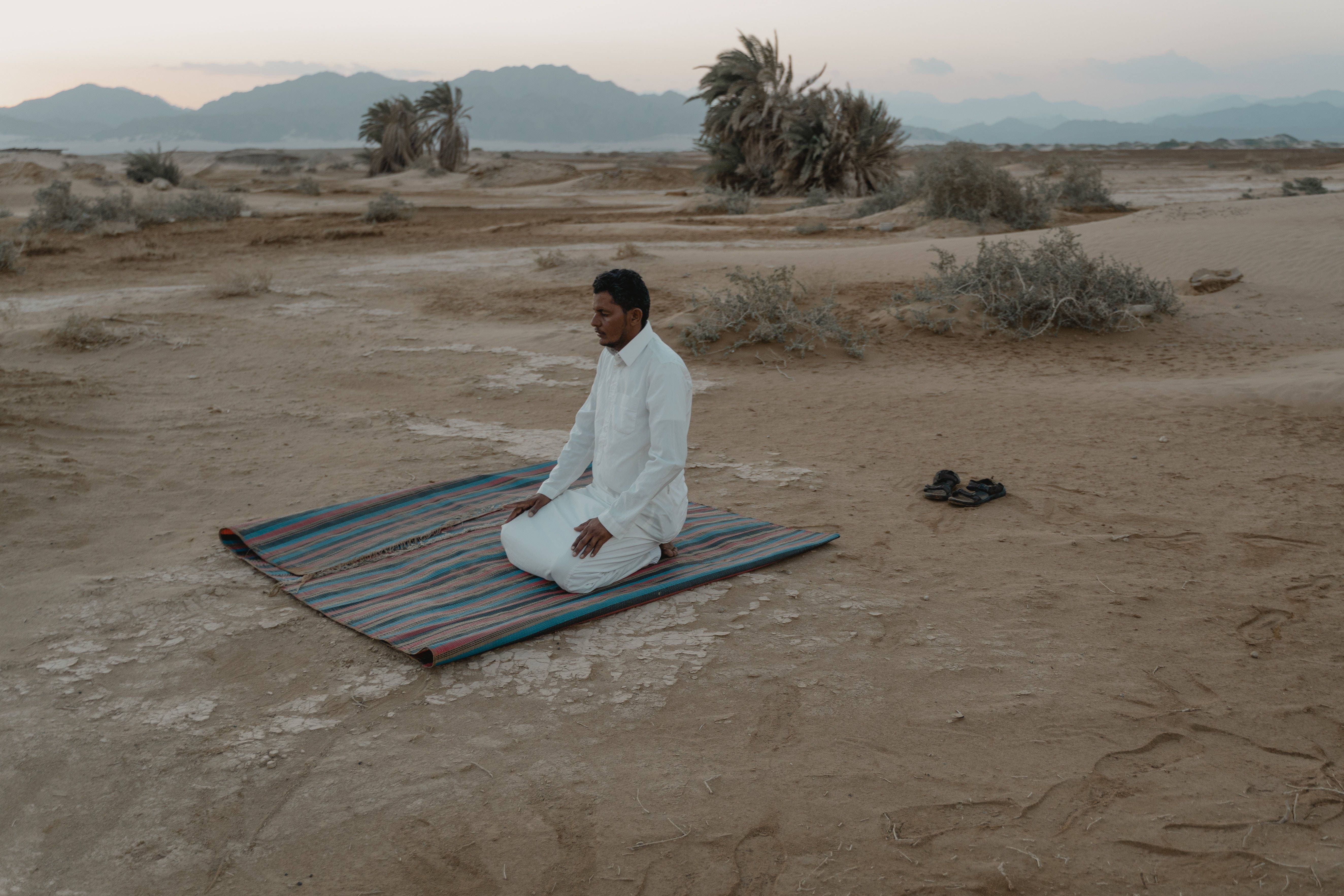 Best Islamic 桌布 壁紙 — 一名身穿白色祈禱服的男人跪在沙漠景觀中的條紋地毯上，黃昏時分，遠處有山脈和棕櫚樹。