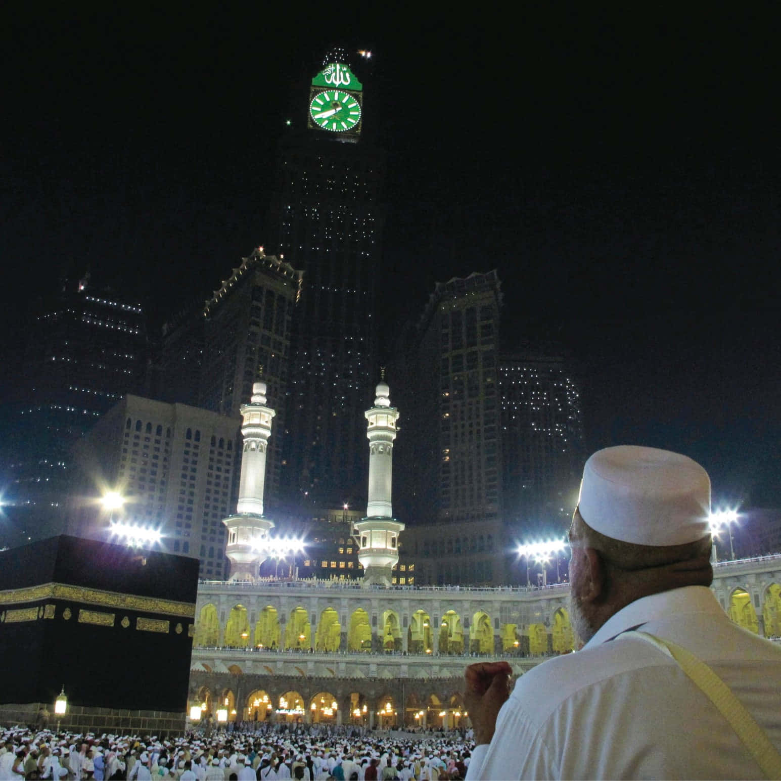 Mecca Mosque 桌布 壁紙 — 一名身穿白色祈禱服的男子抬頭望向夜晚在卡巴天房上方發出綠光的阿布拉吉阿爾拜特鐘樓。