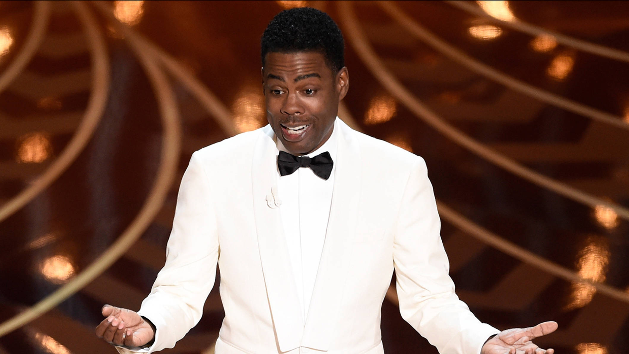 Chris Rock 桌布 壁紙 — 一名身穿白色禮服、繫黑色領結的男子站在舞台上，雙臂張開，背景是金色弧形。