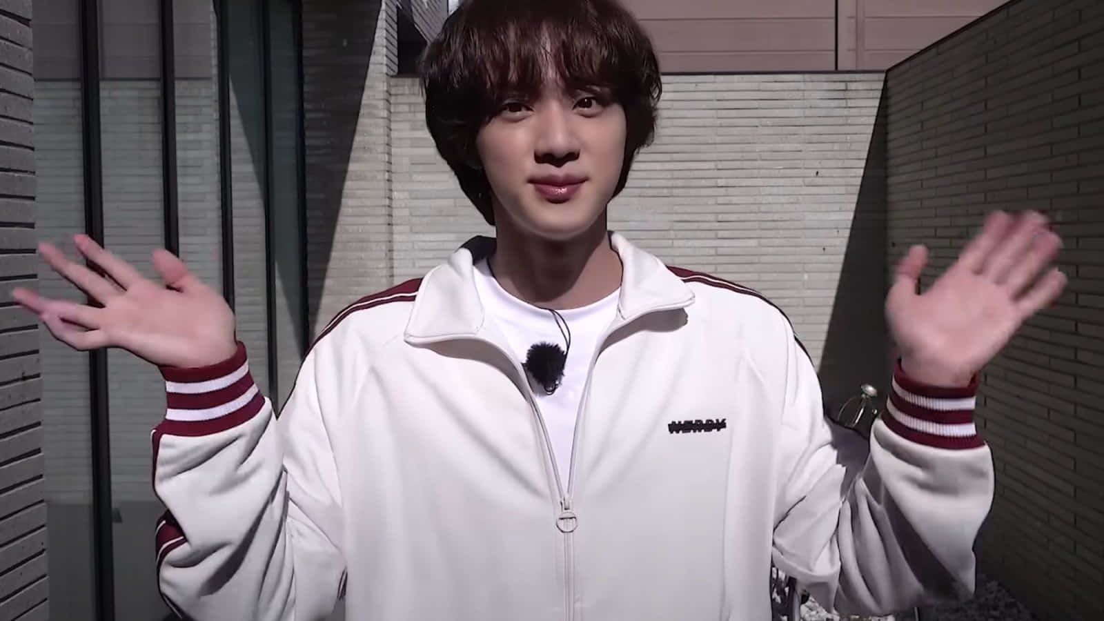 Jin 桌布 壁紙 — 一名身穿白色紅色條紋運動外套的年輕男子，在現代磚牆背景下愉快地揮手。